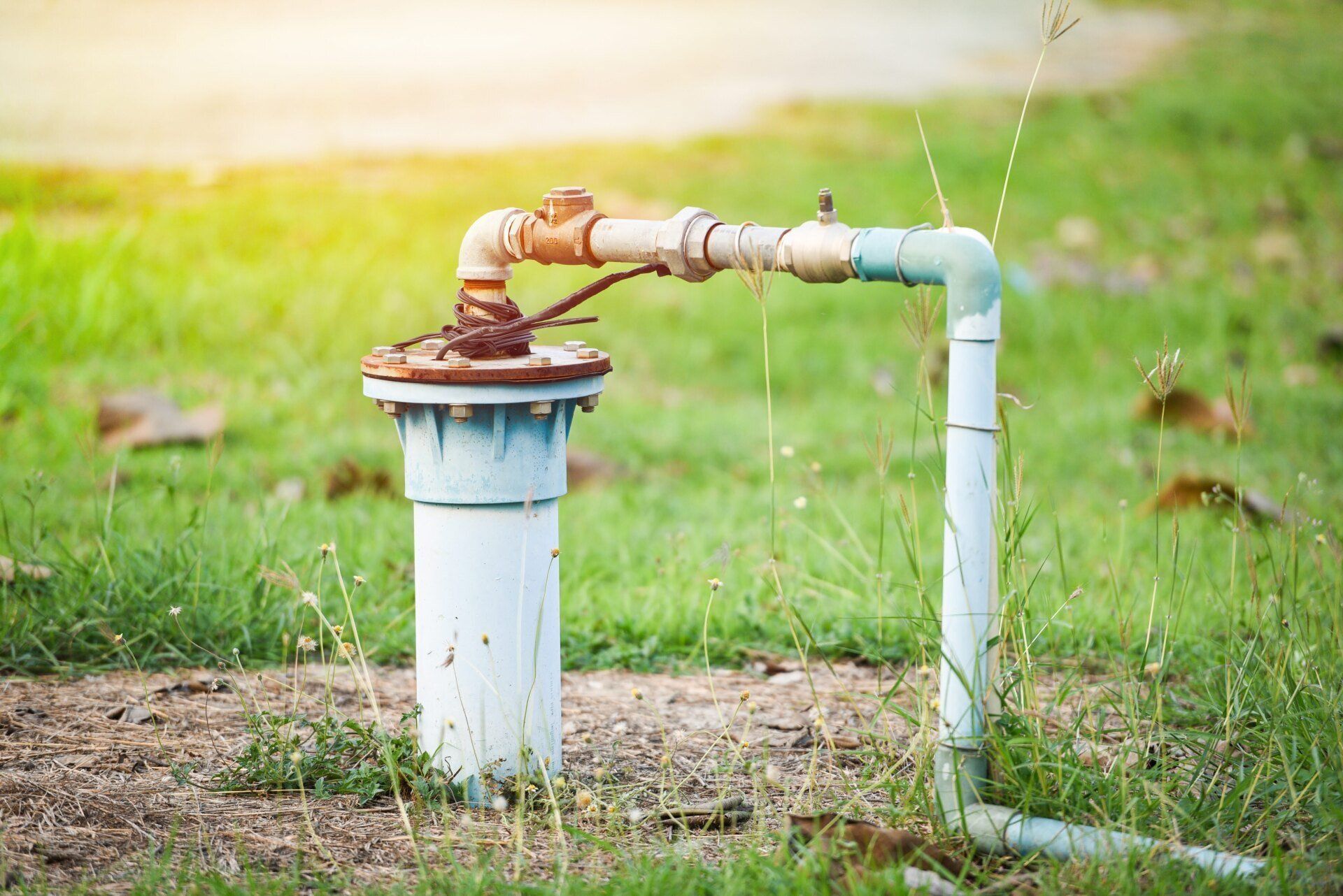 A light blue vertical groundwater pipe assembly with a metal elbow and horizontal pipe fitting, set in a grassy field.