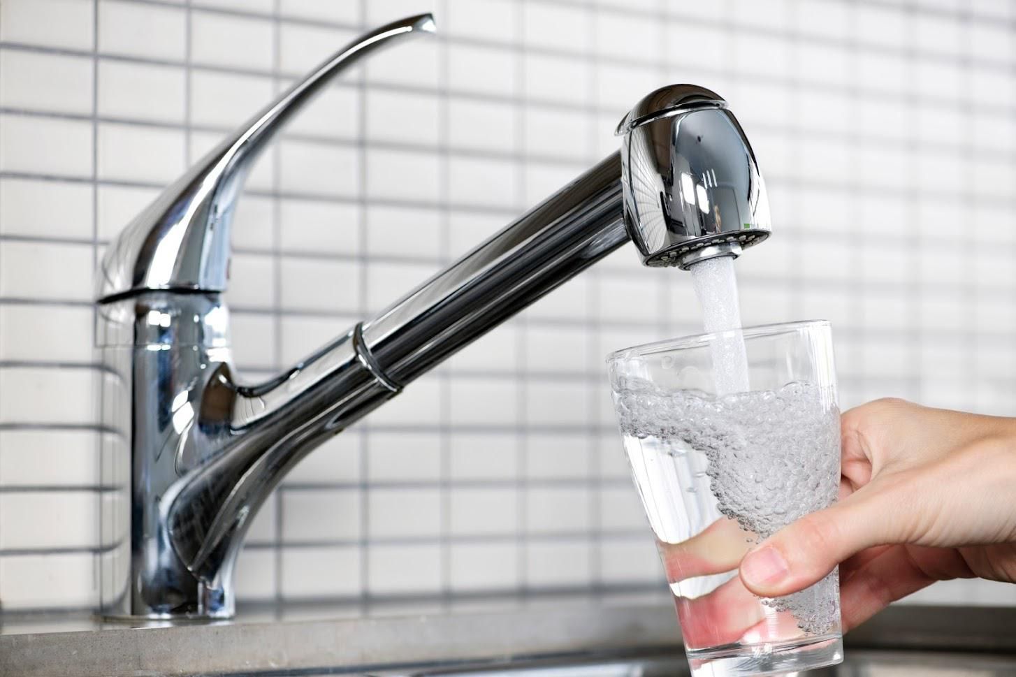 A chrome kitchen faucet pours water into a clear glass held by a hand in front of a white tiled backsplash.