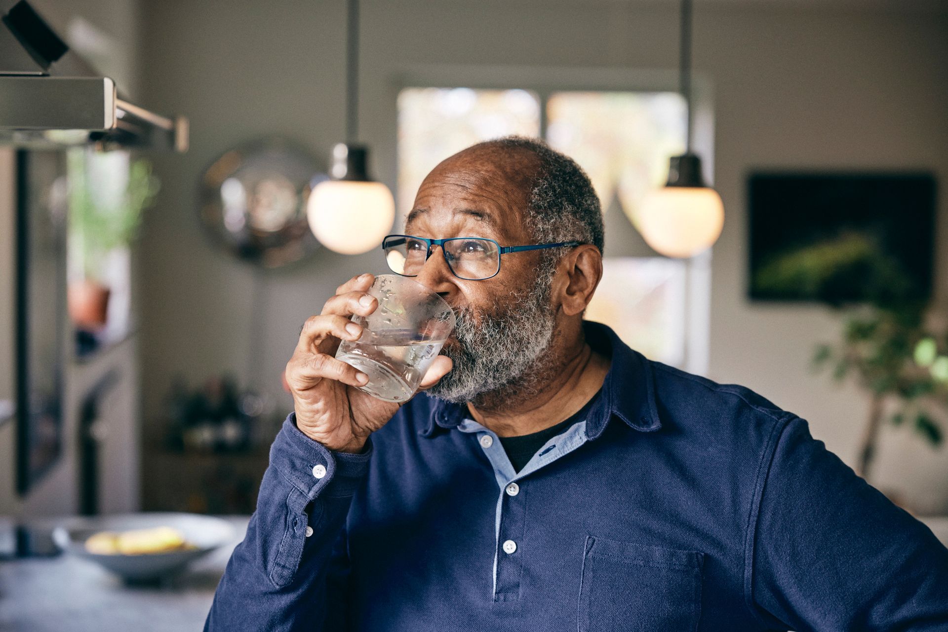 An individual with a gray beard and glasses holds a clear glass of water to their mouth in a softly lit, modern home.