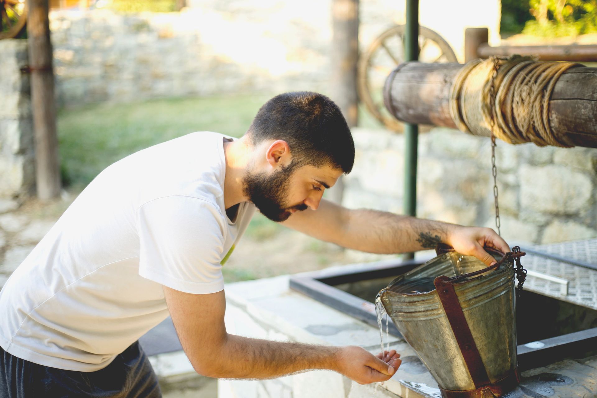 A man with a beard leans over a stone well, catching water pouring from a metal bucket into his cupped hands.
