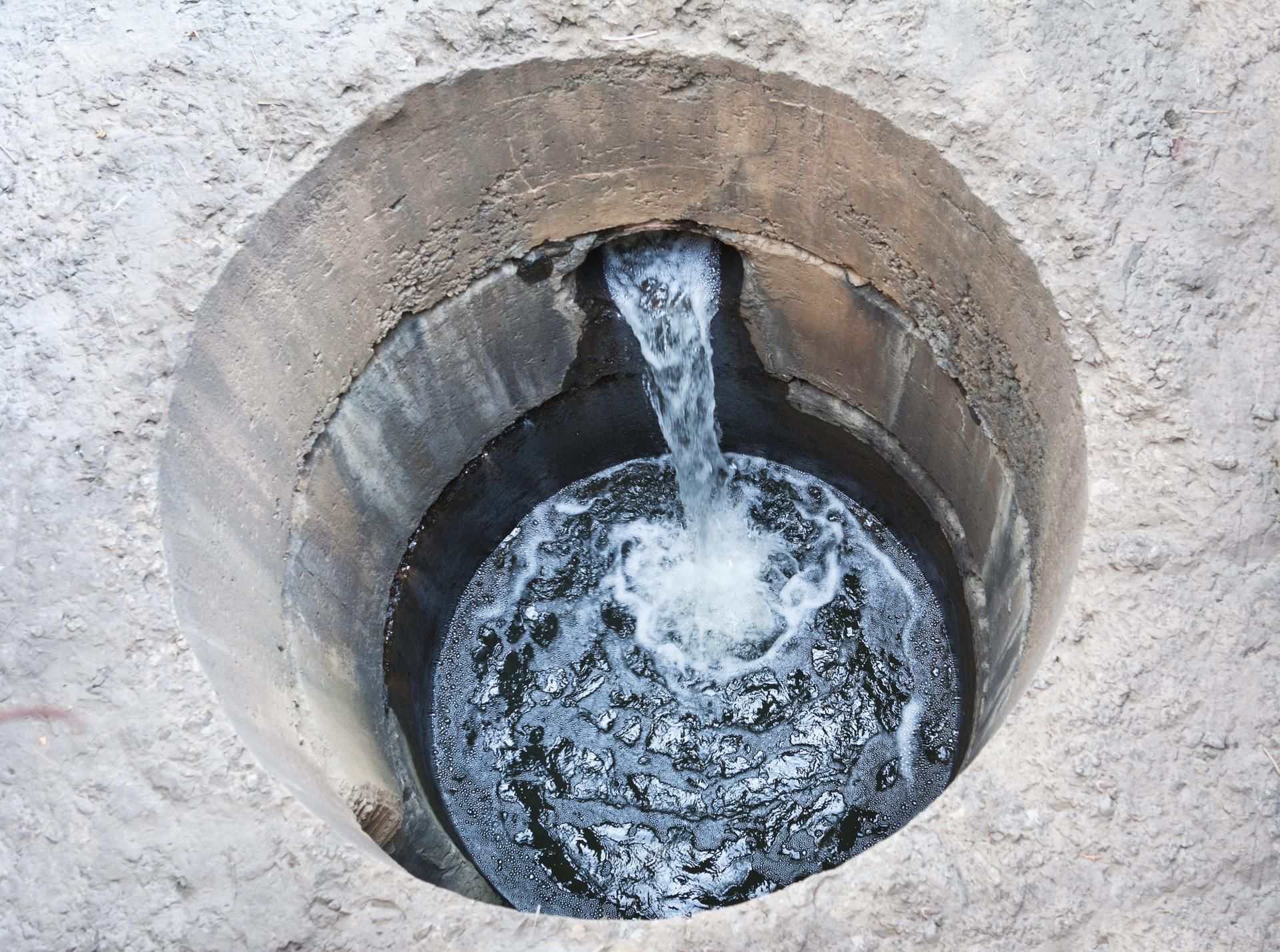 Water flows from a pipe into a round, concrete-lined well or basin set into a textured, light-colored ground surface.