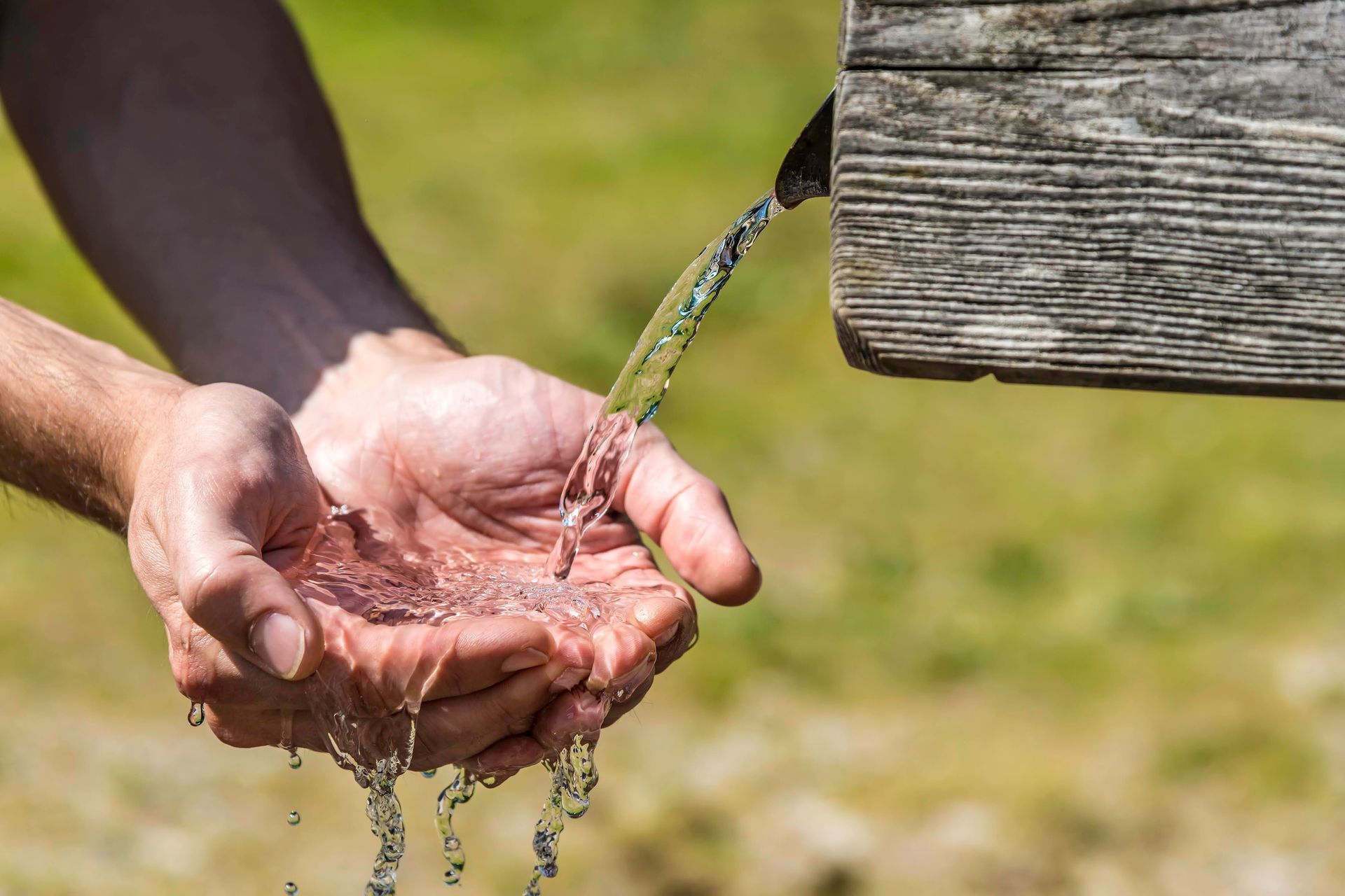 Clear water flowing from a rustic wooden spout into a pair of cupped hands.