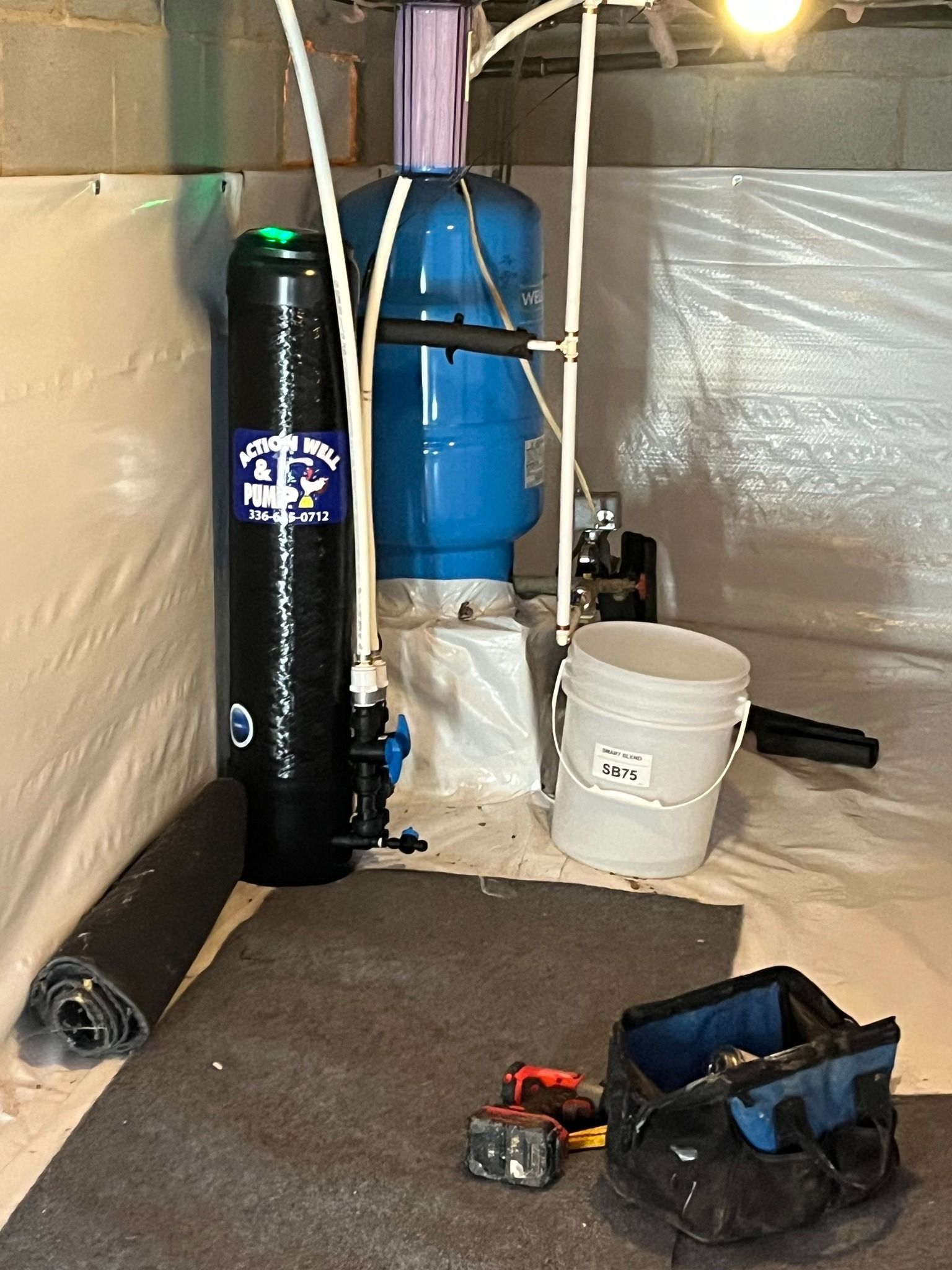 A water filtration system and blue pressure tank stand in a basement corner near a bucket and a tool bag on the floor.