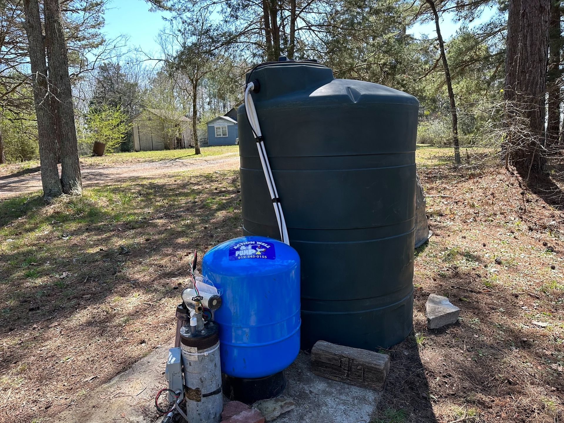 A large dark green water storage tank next to a blue pressure tank on a concrete pad in a wooded outdoor setting.