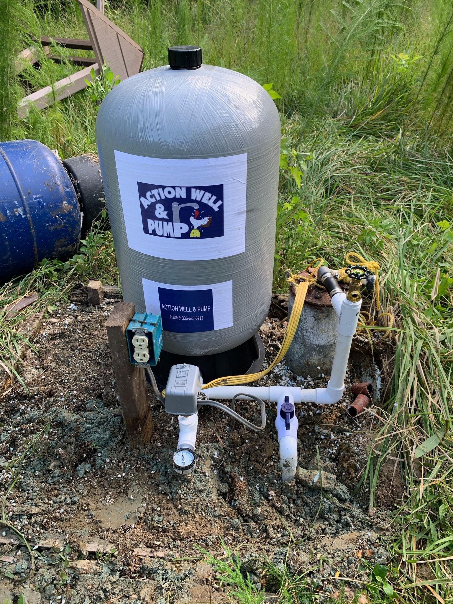 A grey well pressure tank with a pump label, located in a grassy outdoor area, connected to pipes and electrical outlets.