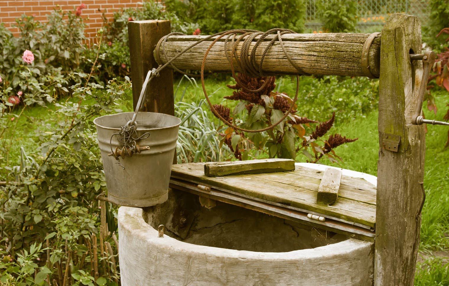 A rustic stone well with a metal bucket hanging from a weathered wooden beam in a garden.