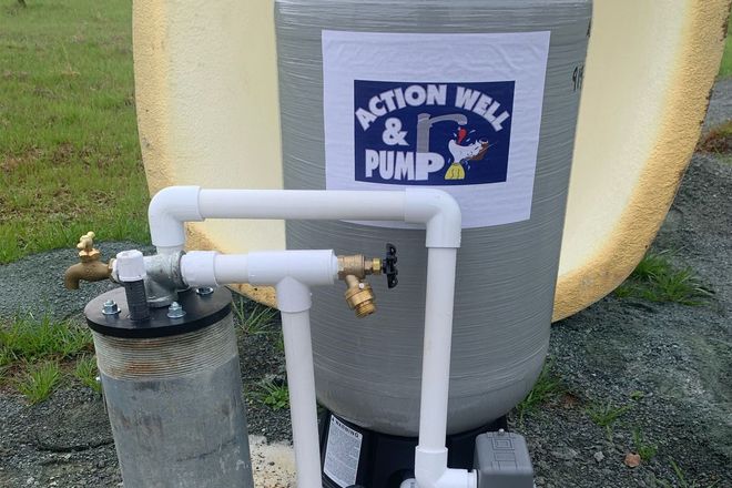 Gray tank with white PVC pipes and a black top, labeled “Action Well & Pump” outdoors on gravel.