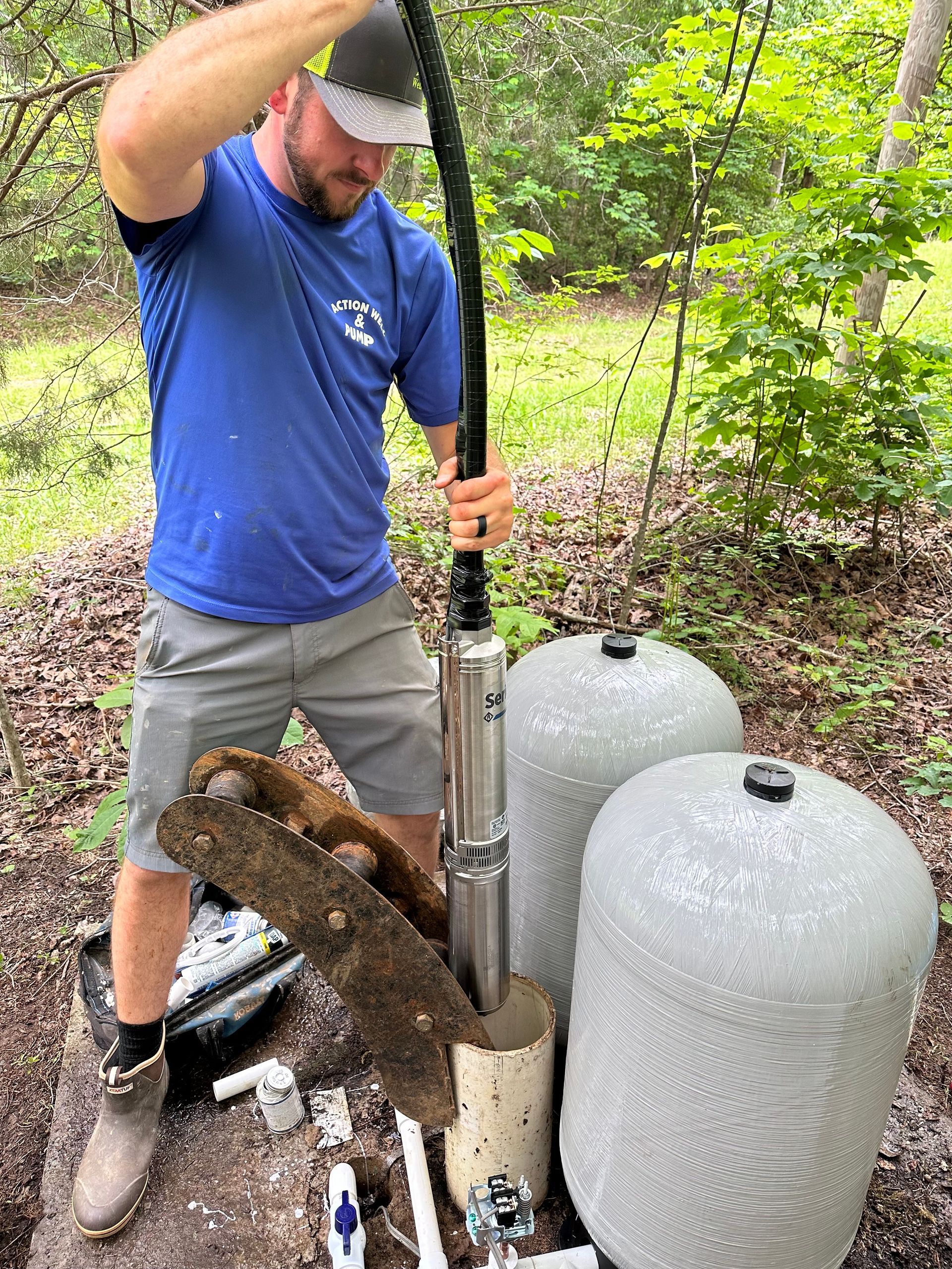 A person in a blue shirt lowers a long, cylindrical well pump into a pipe outdoors next to two large grey water tanks.