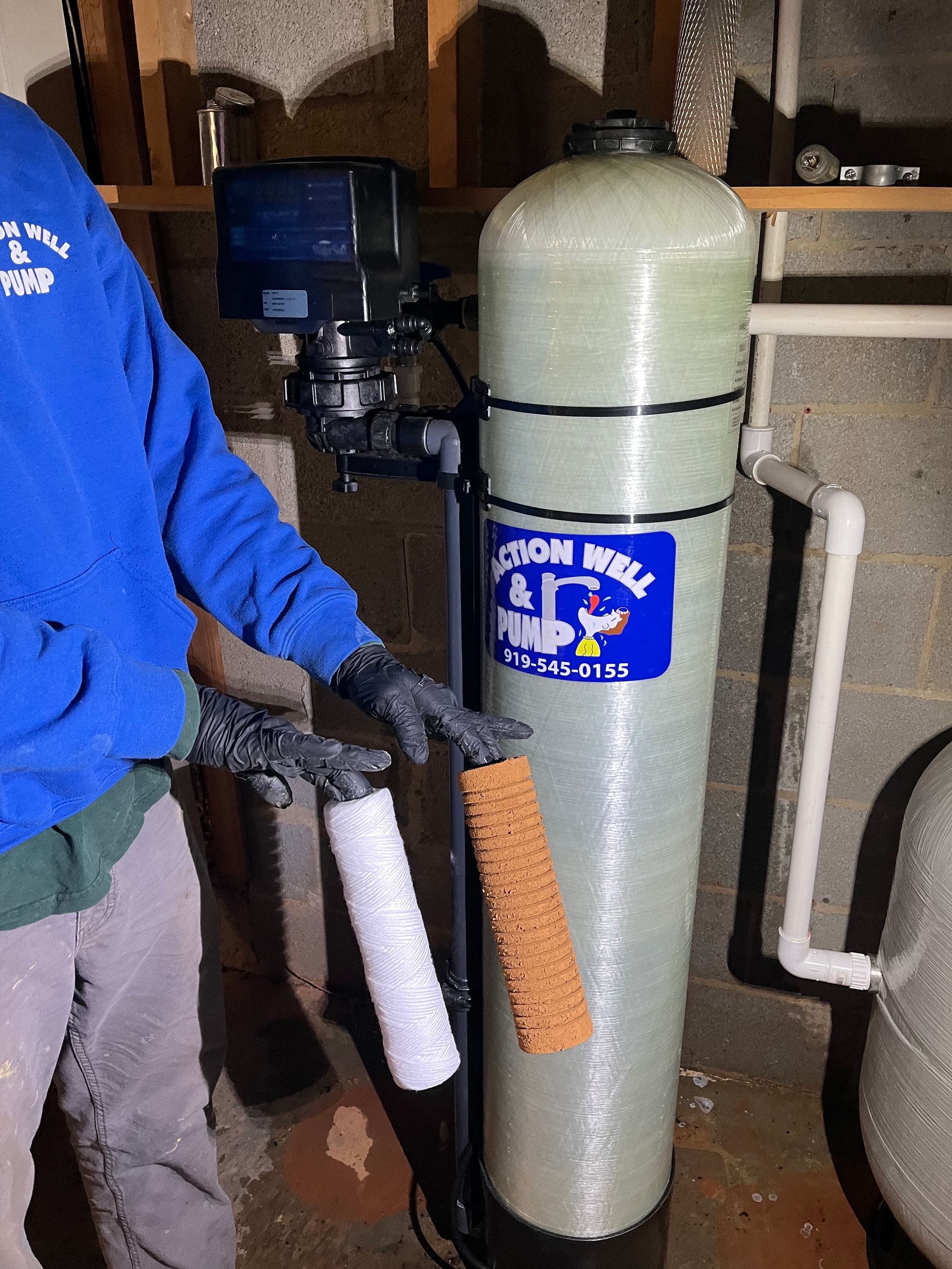 A person in a blue hoodie holds a clean white water filter next to a dirty, orange-stained filter near a well water tank.