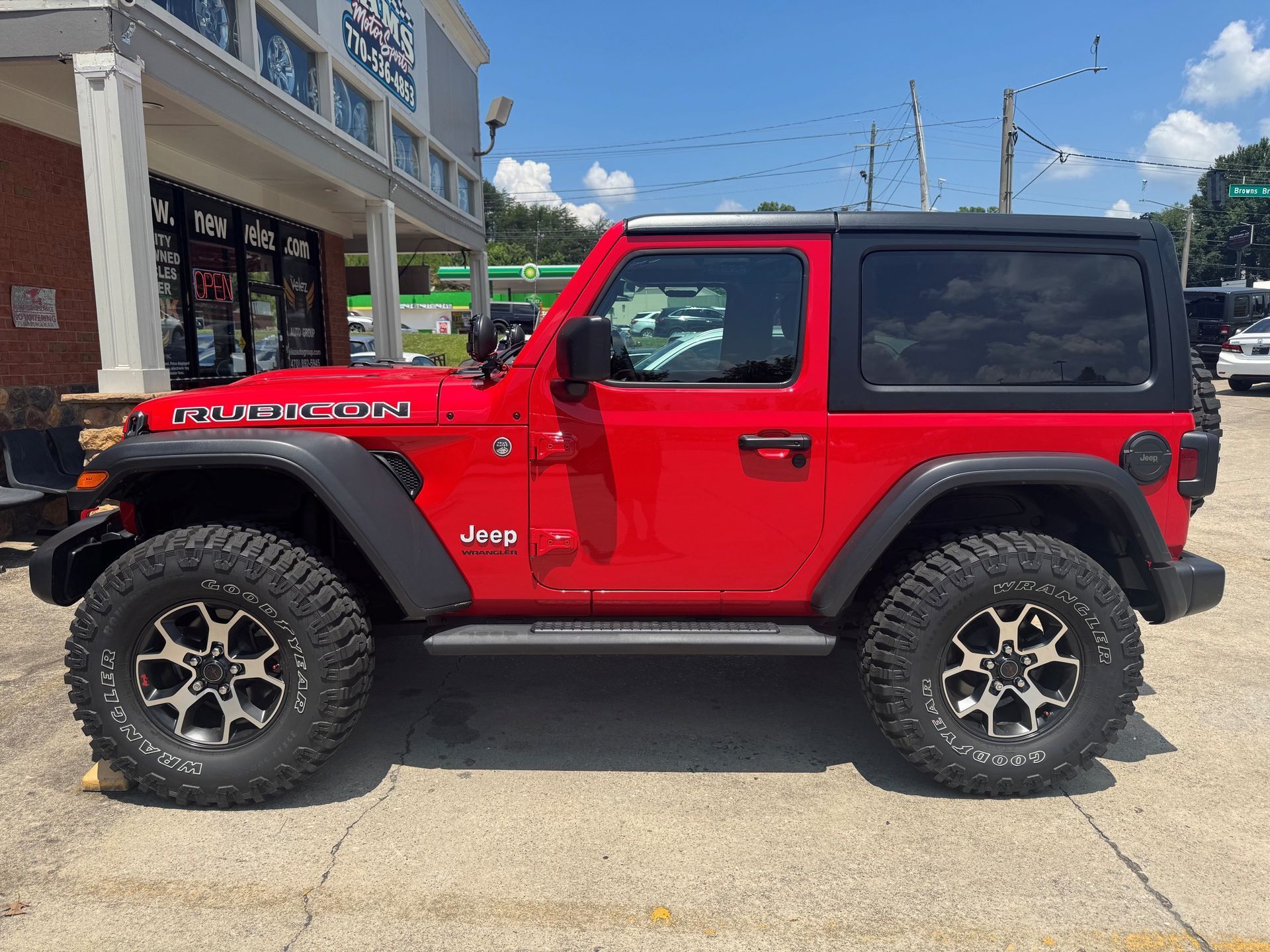 Red two-door Jeep Wrangler parked outside a storefront, with black hardtop and off-road tires.