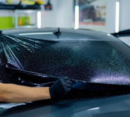 Gloved hands apply a black vinyl wrap to a car hood in a workshop