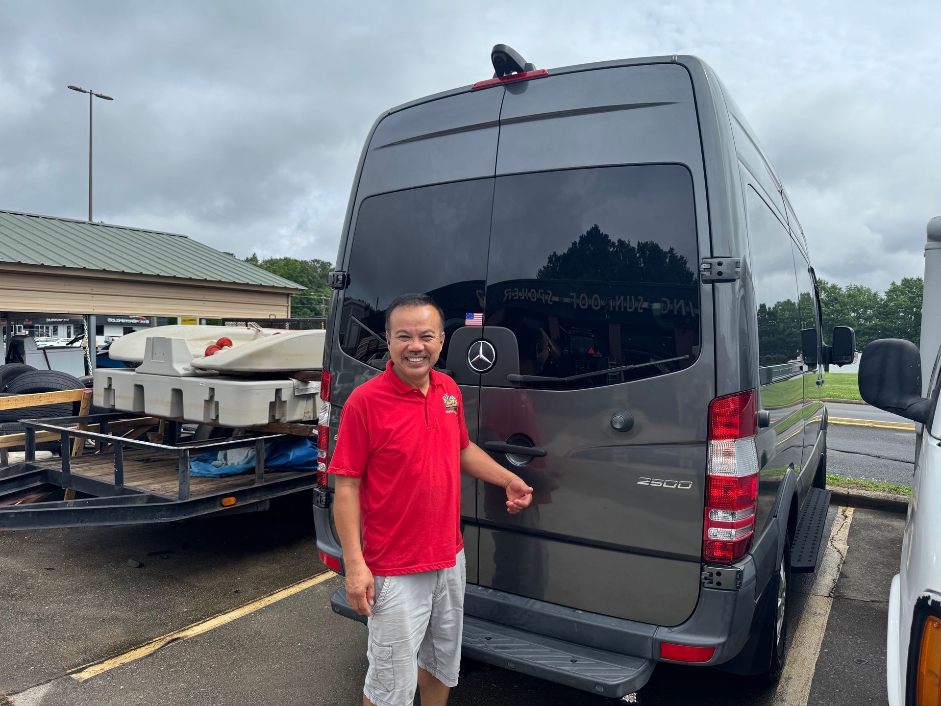 Man in red shirt stands beside a dark van in a parking lot under cloudy skies