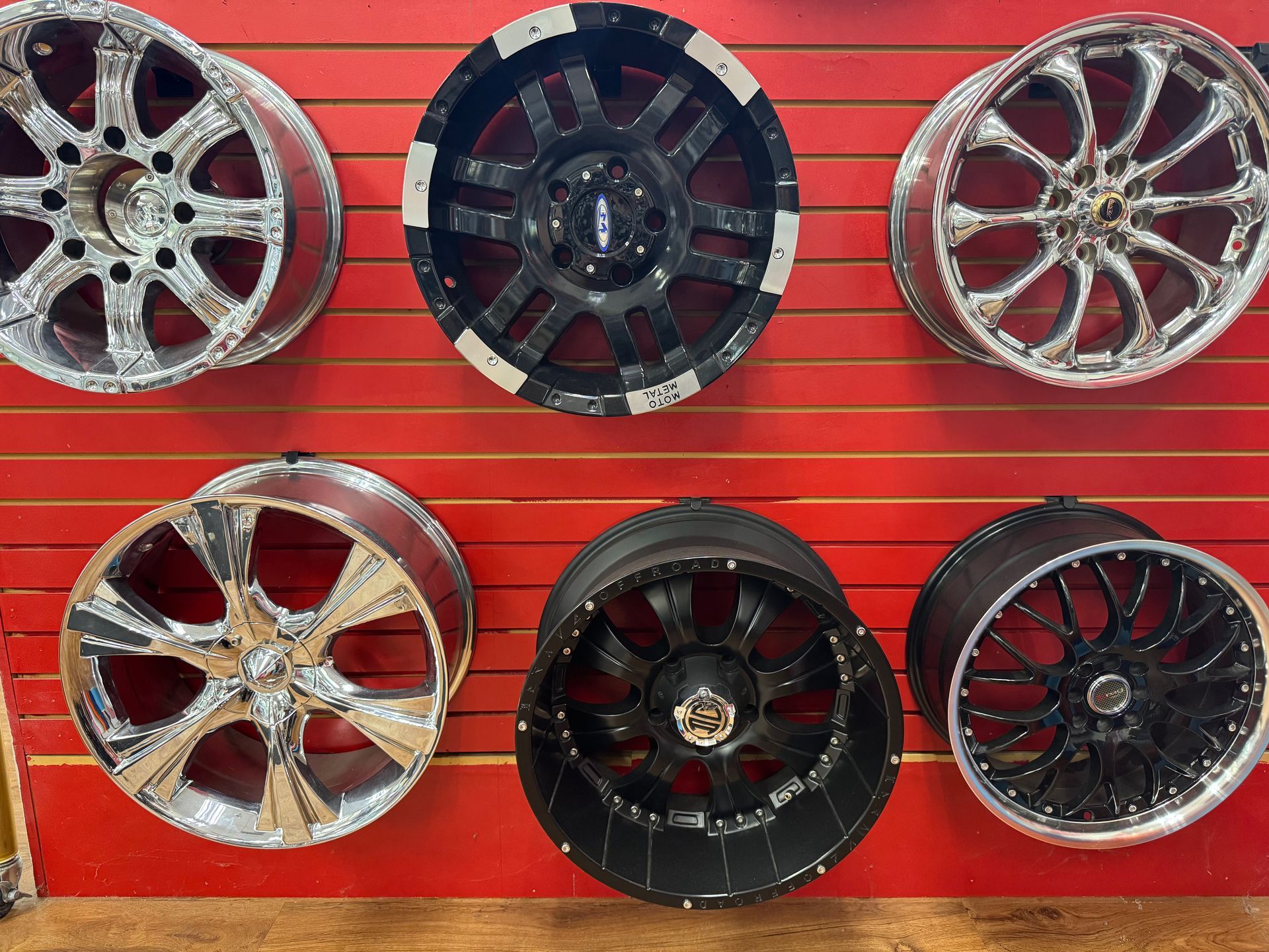 Six decorative car rims displayed on a red slatwall, in silver, black, and chrome finishes.