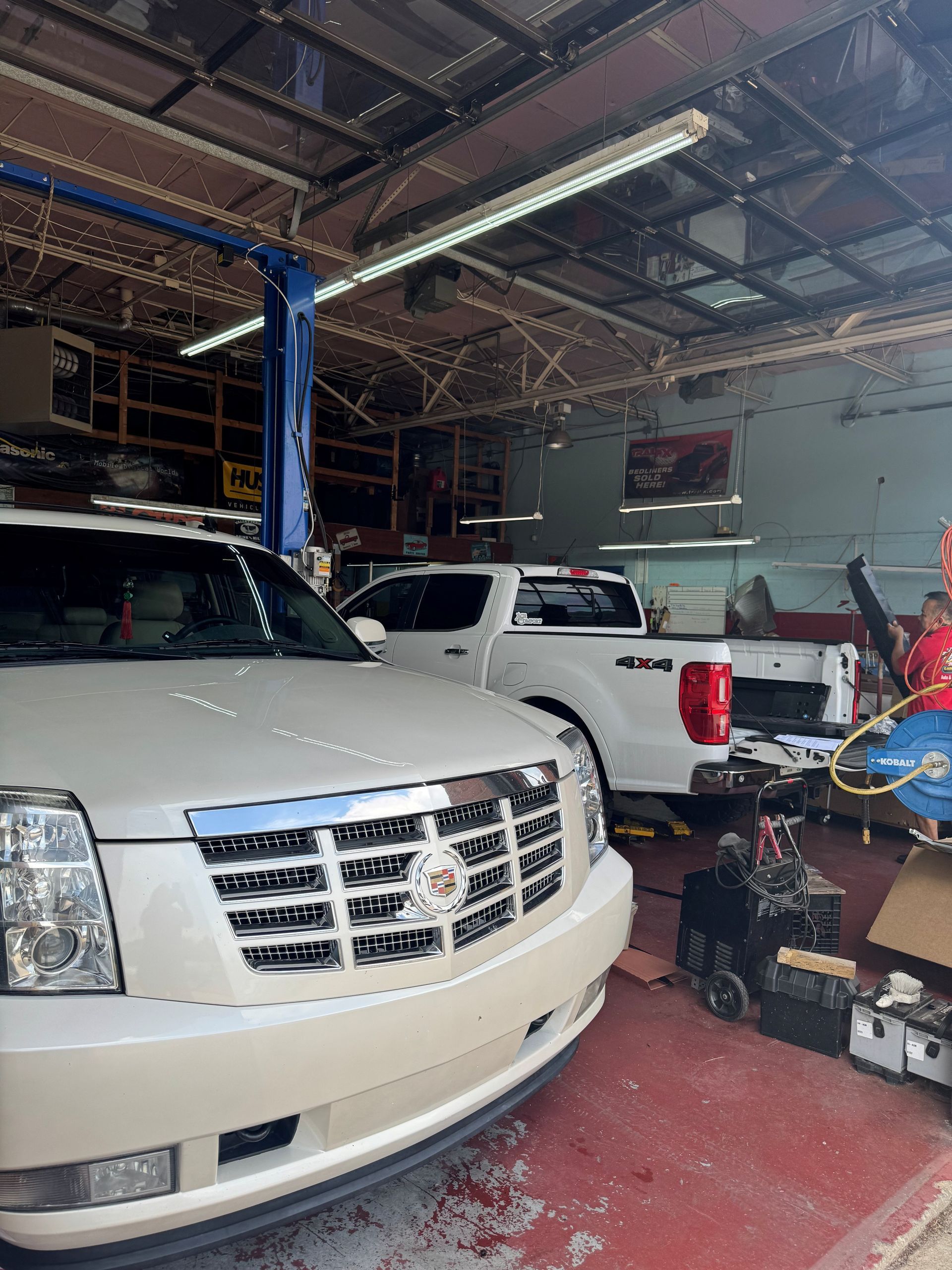 White SUV in a cluttered auto repair garage with tools, equipment, and another white pickup truck nearby