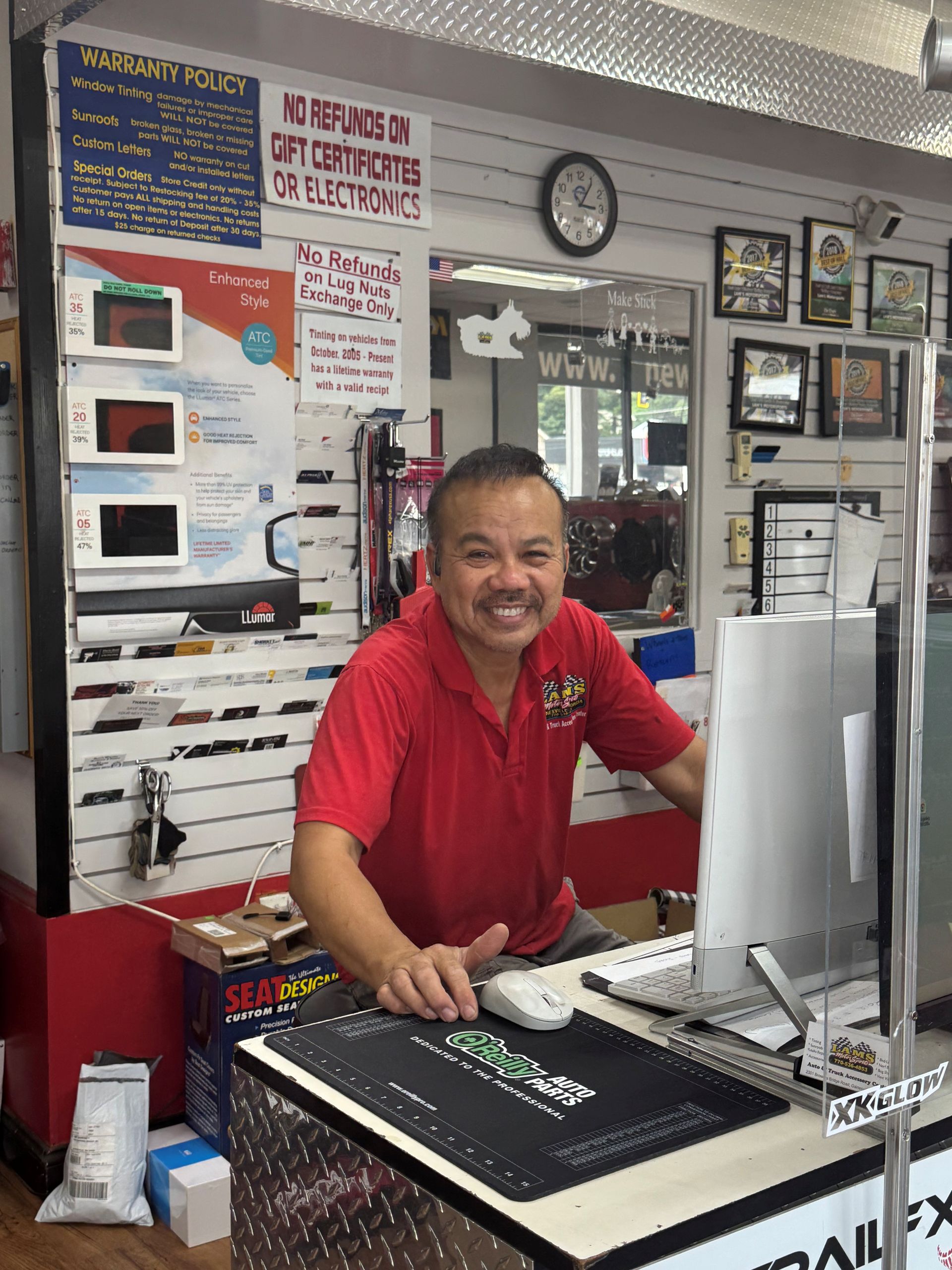 Store clerk in a red shirt smiling behind a counter with a computer in a small retail shop.
