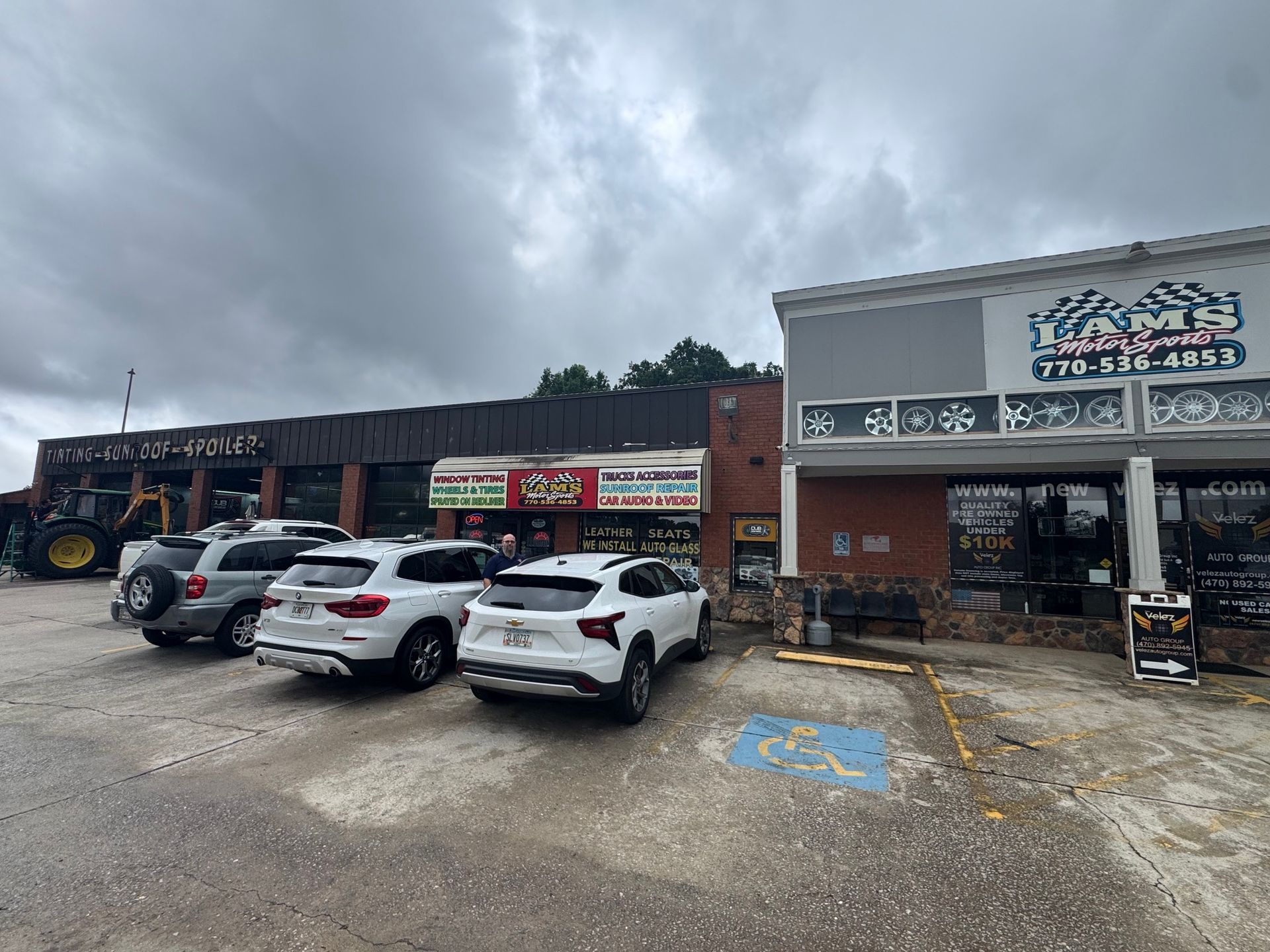 Strip mall storefronts with parked cars under a cloudy sky