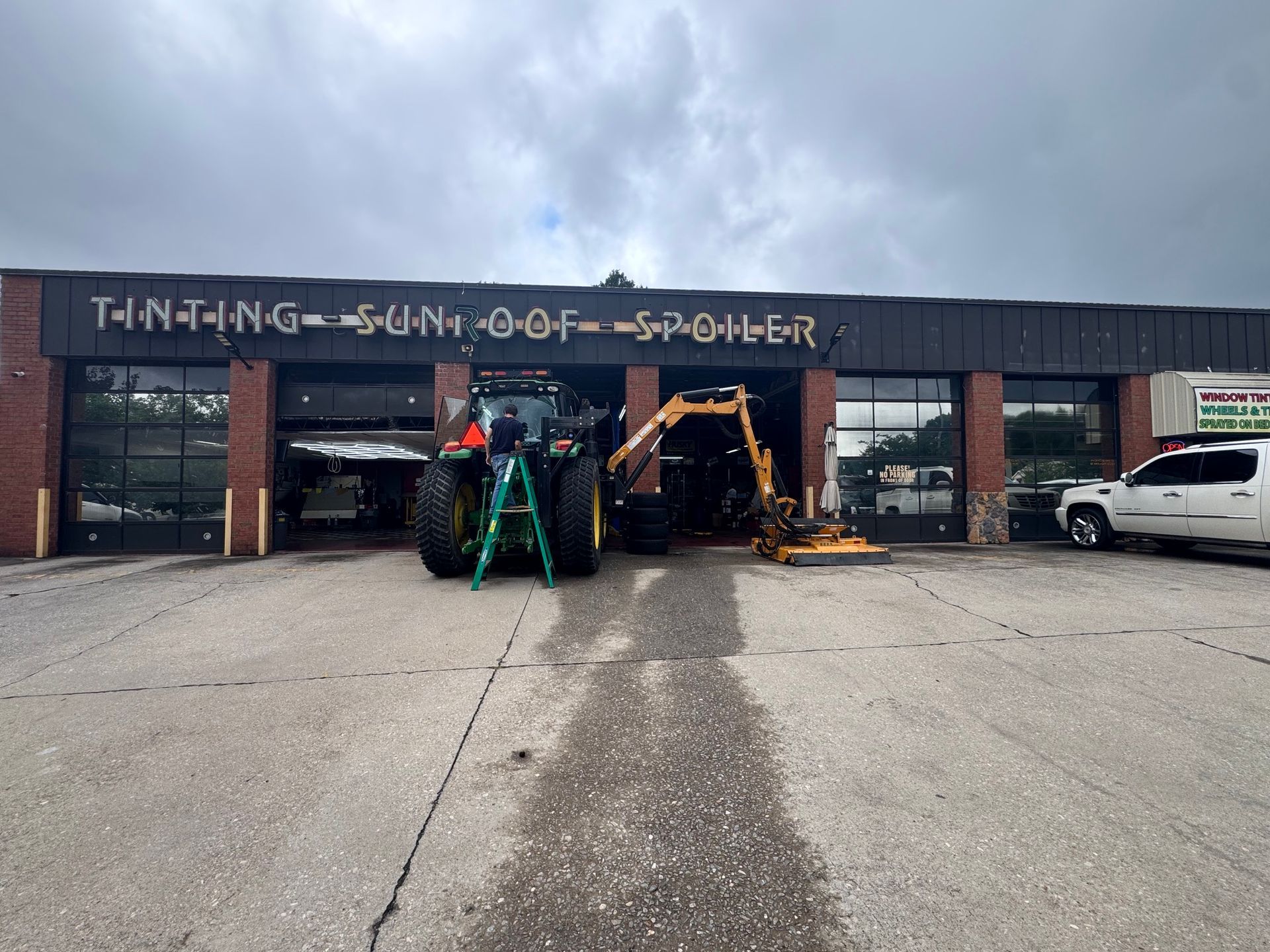 Construction vehicles parked in an open garage with a white SUV beside them under a cloudy sky
