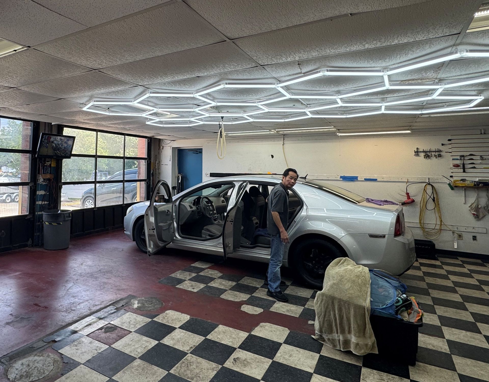 Silver car in a garage with doors open and a person standing beside it under hexagonal ceiling lights