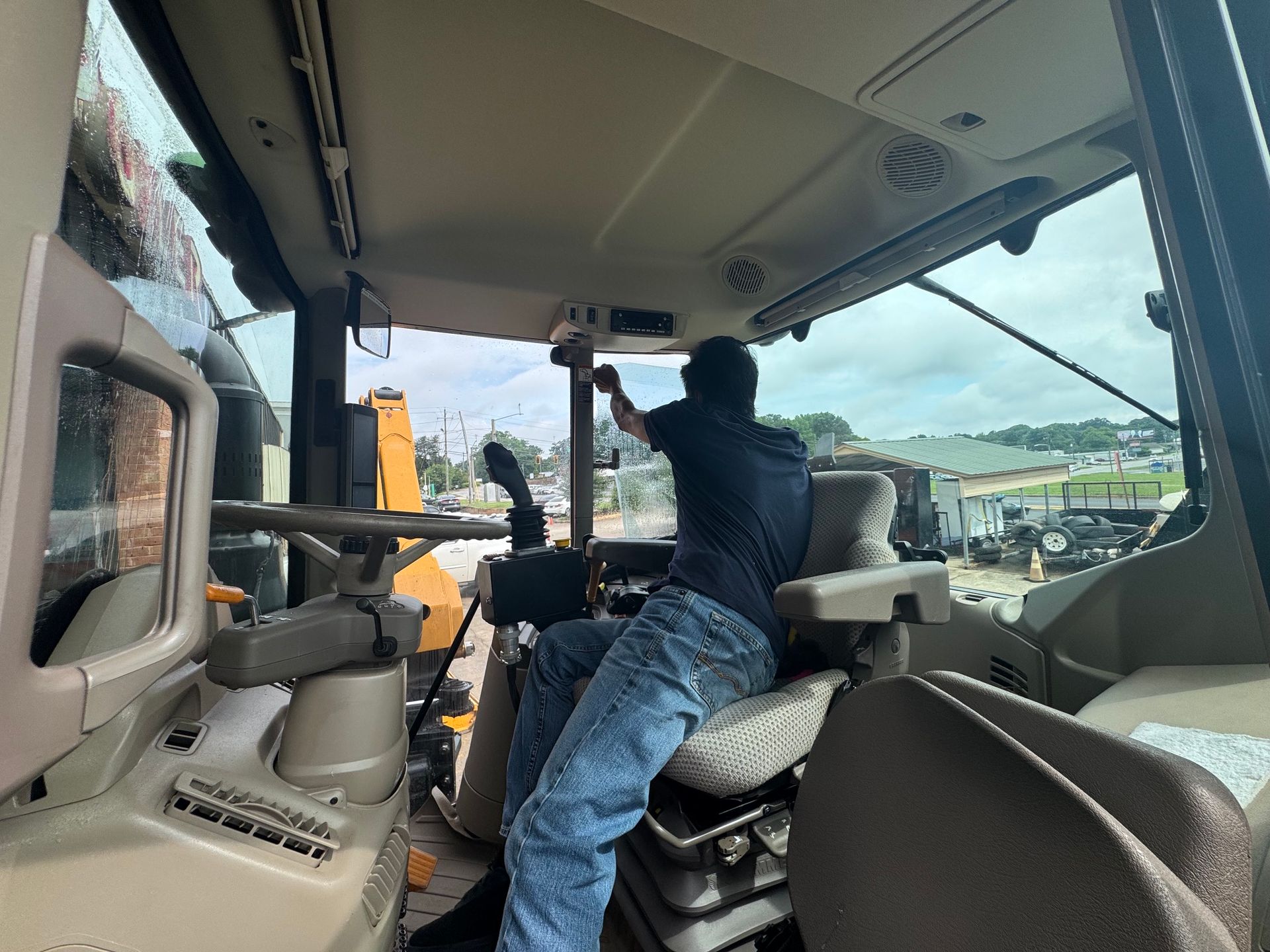Person seated in a vehicle cab, reaching up to adjust the overhead controls, with a construction site outside.