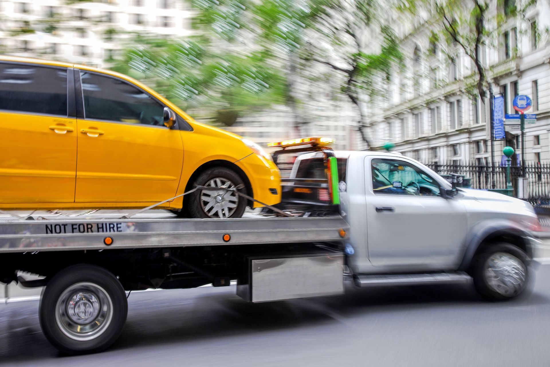 Tow truck hauling a yellow car on a city street, with buildings blurred in the background