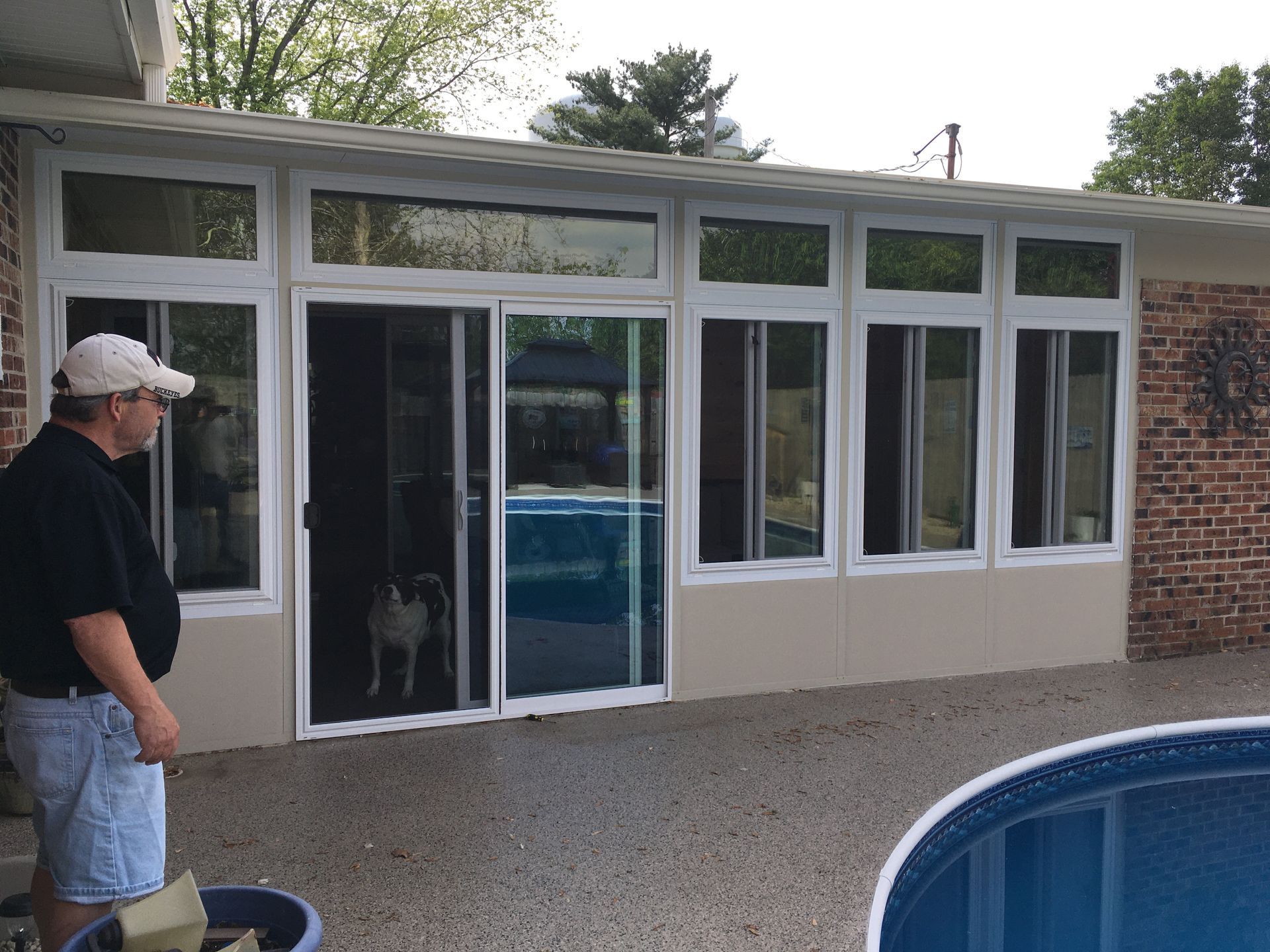 Man and dog by a pool, looking at a sunroom with sliding glass doors. White frames, beige panels, brick building.