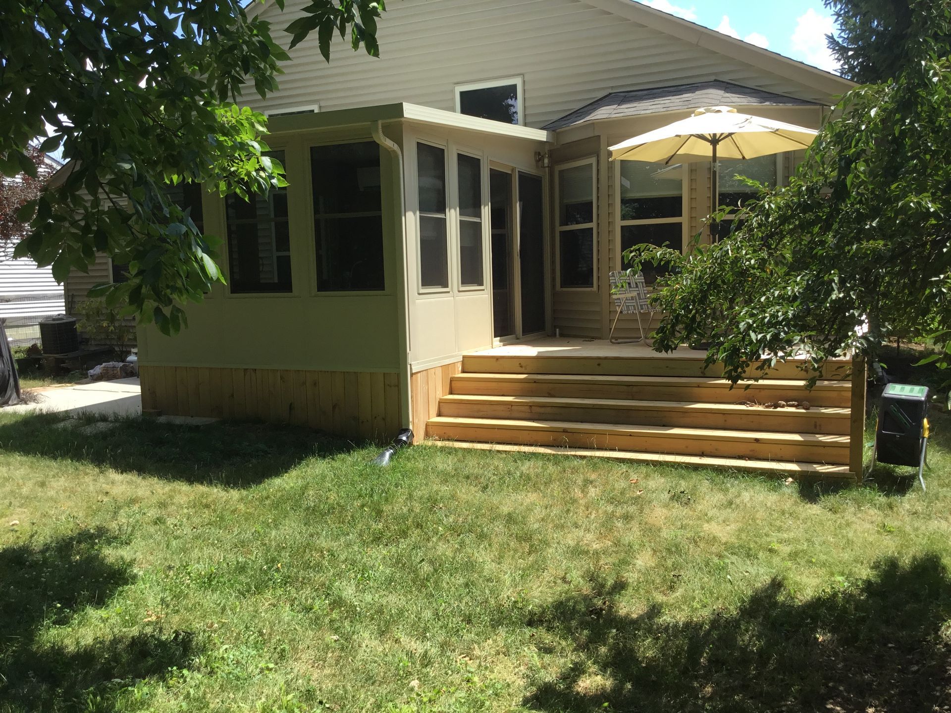 Screened porch with steps, attached to a light-colored house. Deck has an umbrella, surrounded by grass and trees.