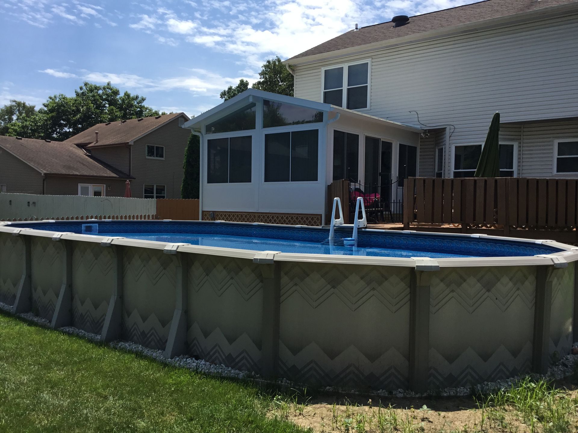 Above-ground pool in backyard with attached sunroom and residential housing in background.