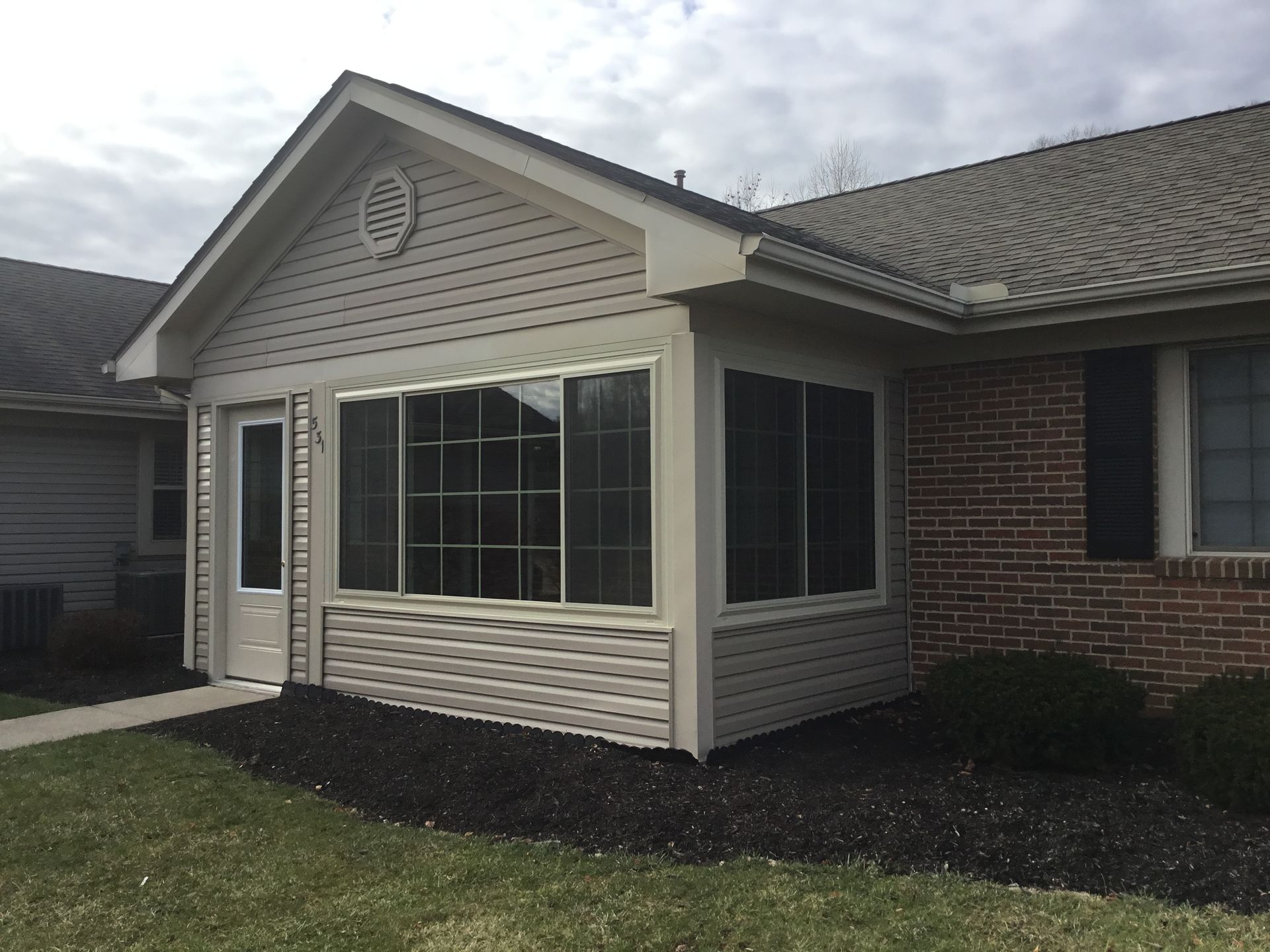Exterior view of a beige sunroom with large windows, a door, and a dark mulch bed next to a brick building.