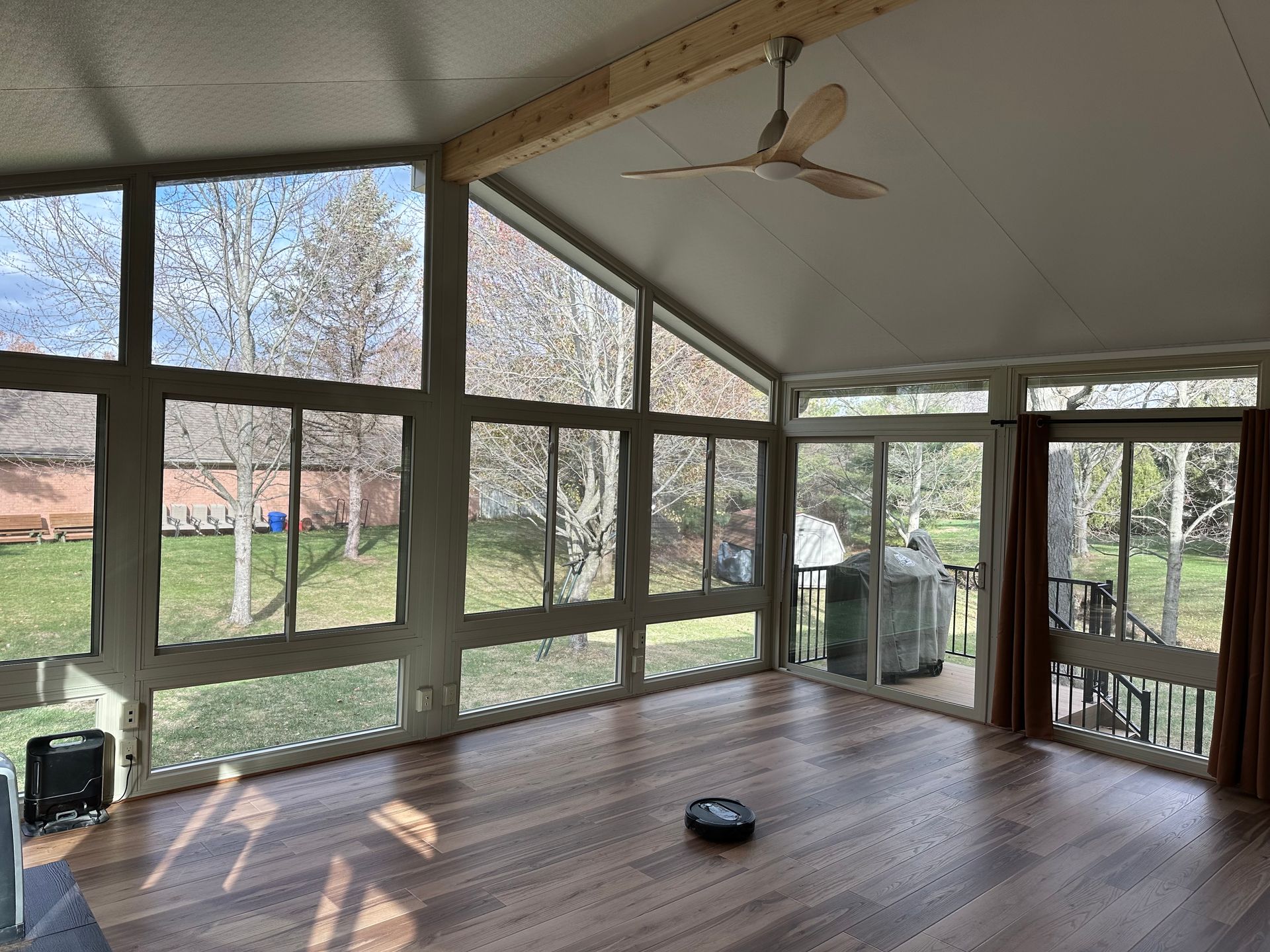 Sunroom with large windows and a ceiling fan; robot vacuum on the hardwood floor; view of trees outside.