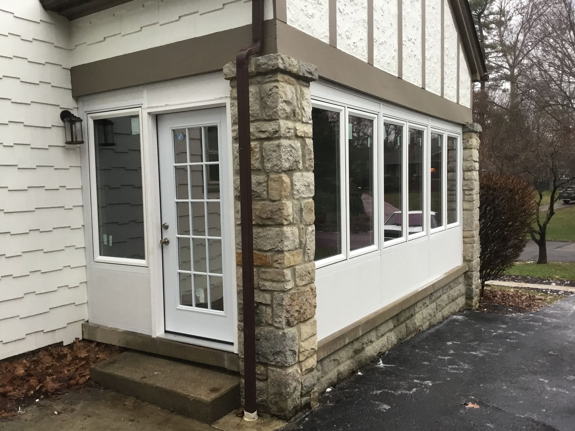 Enclosed porch with white trim, door, and windows. Brown roof trim, stone columns, and asphalt driveway.