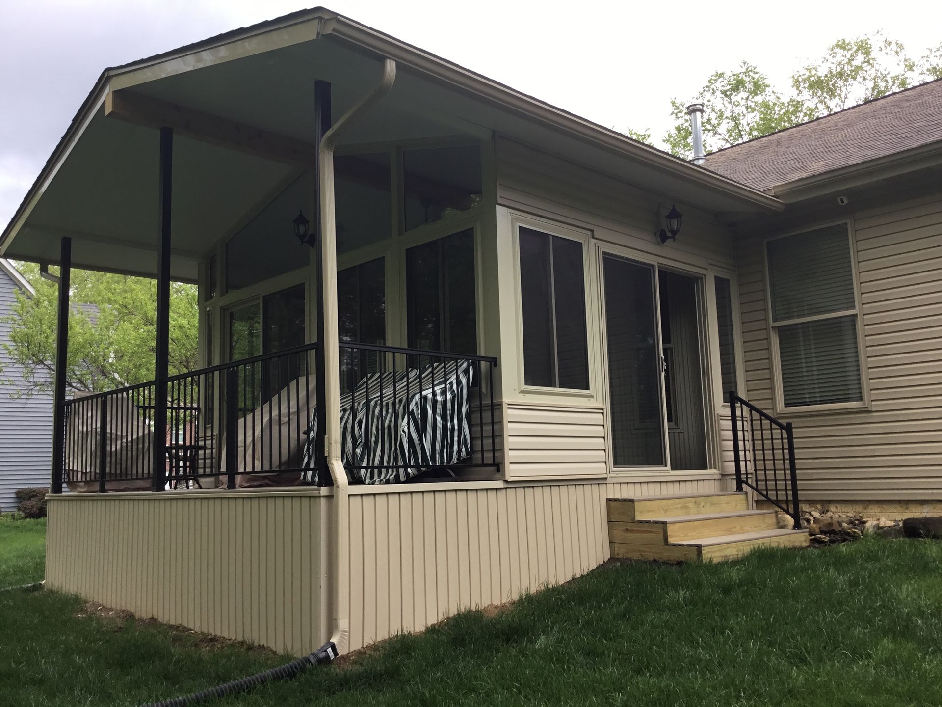 Beige enclosed porch with black railing, steps, and siding, connected to a house with beige siding and green grass.
