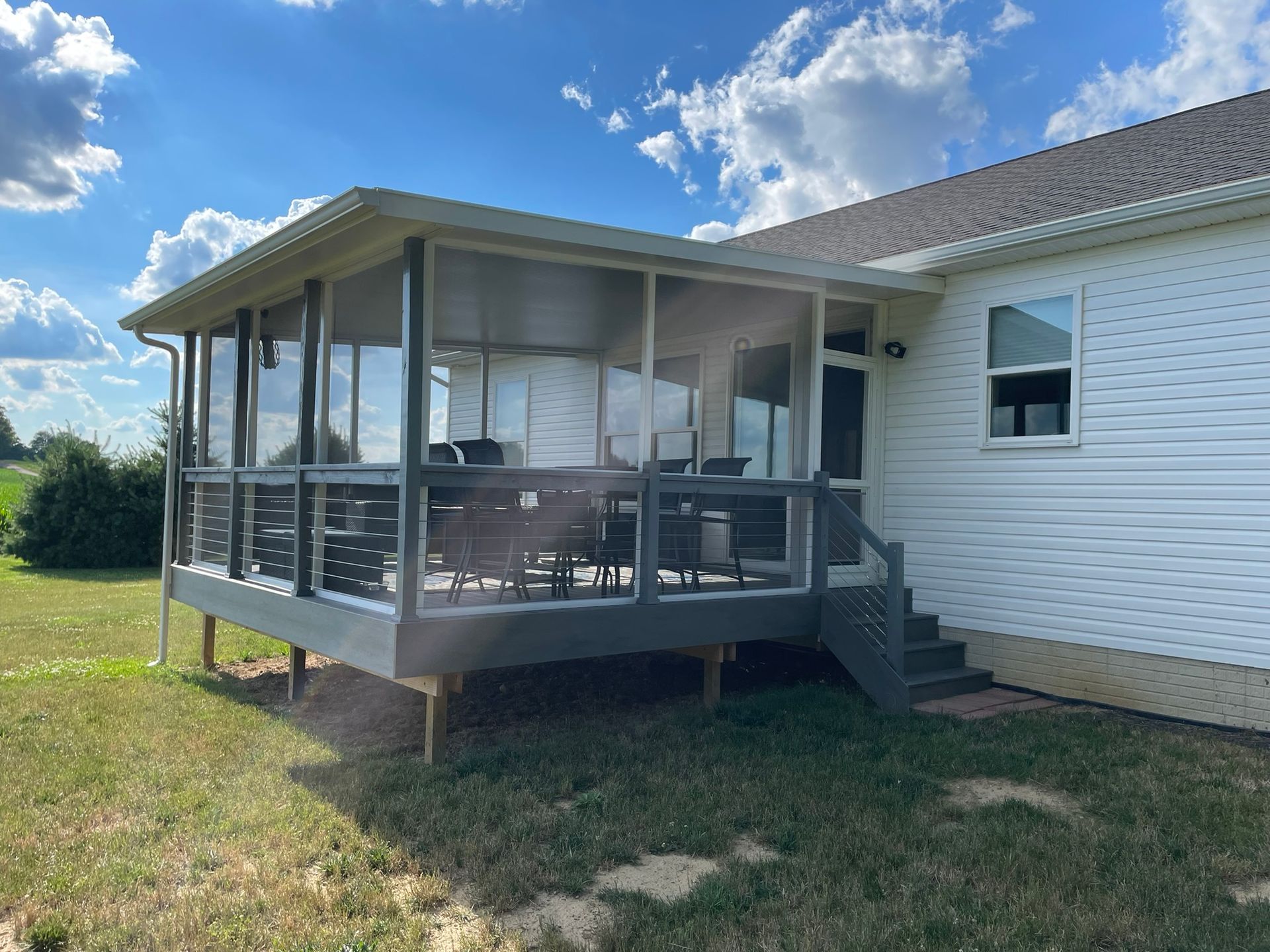 Screened-in porch attached to a white house with outdoor furniture. Green grass and blue sky with clouds in the background.