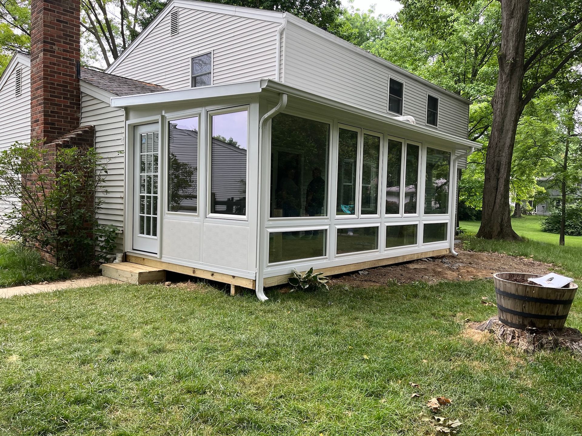 White-framed sunroom addition on a two-story house, with windows, a door, and a wooden deck, set on a grassy lawn.