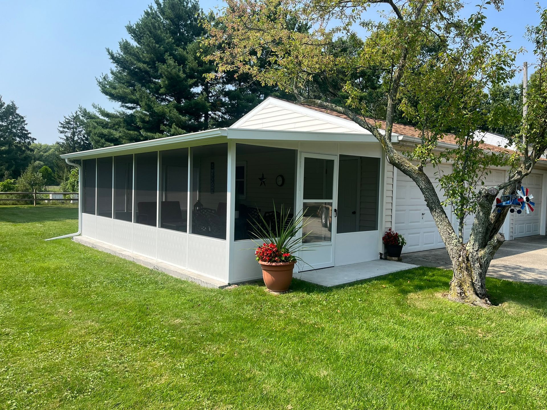 White screened-in porch with a gabled roof, potted flowers, and a grassy yard.
