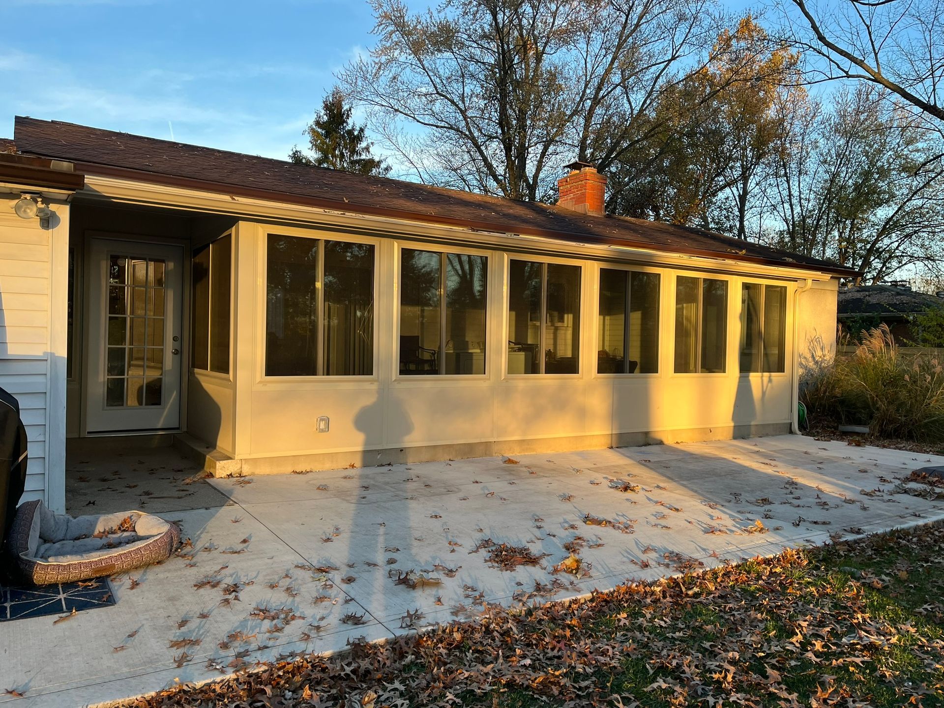 Sunroom with concrete patio, tan walls, brown roof, and several windows. Autumn leaves scattered around.