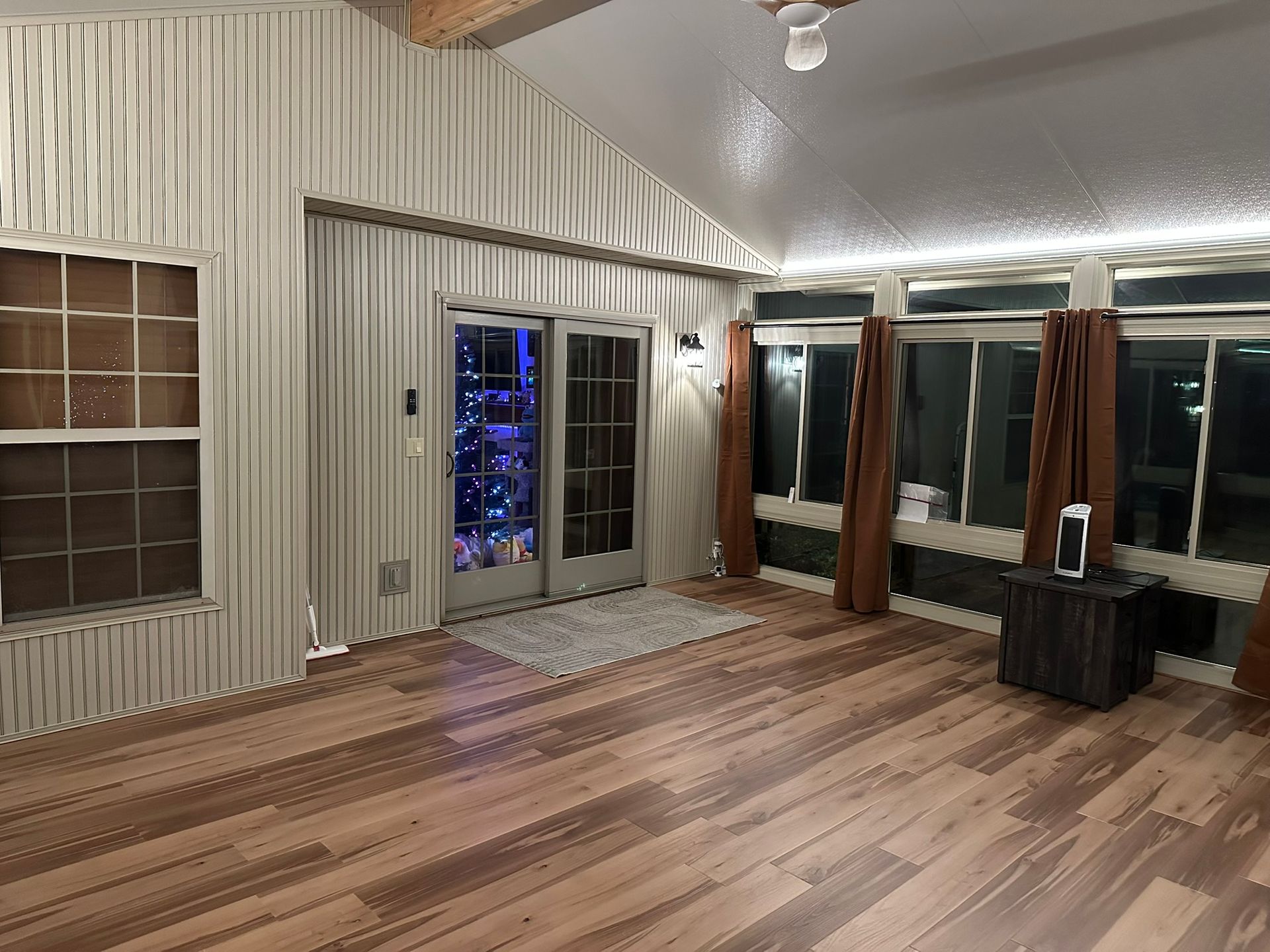 Sunroom with wooden floors, large windows, and brown curtains. A door with holiday lights is visible.