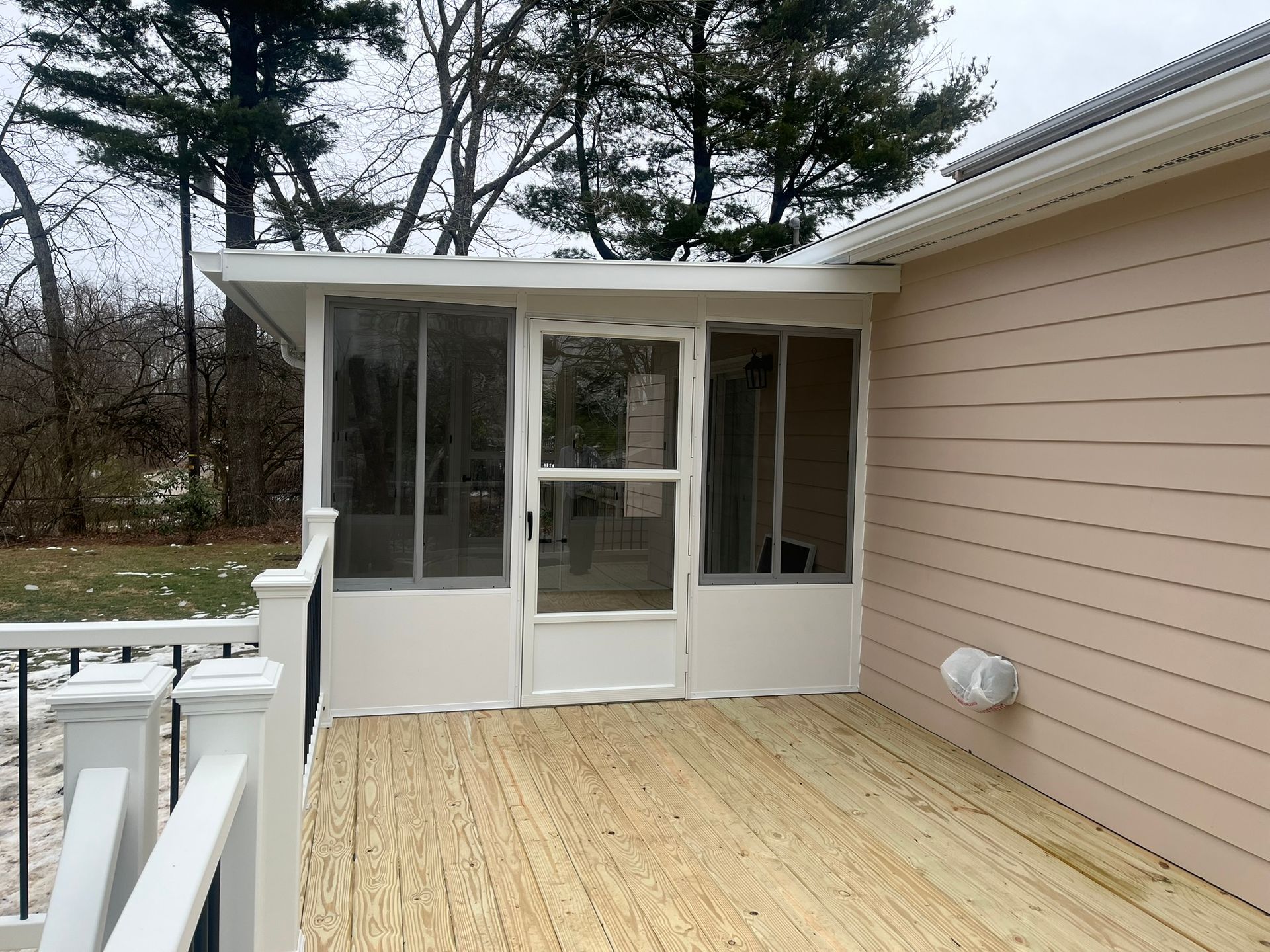 Screened-in porch on a wooden deck, attached to a tan house. White trim, glass and screen windows, trees in the background.