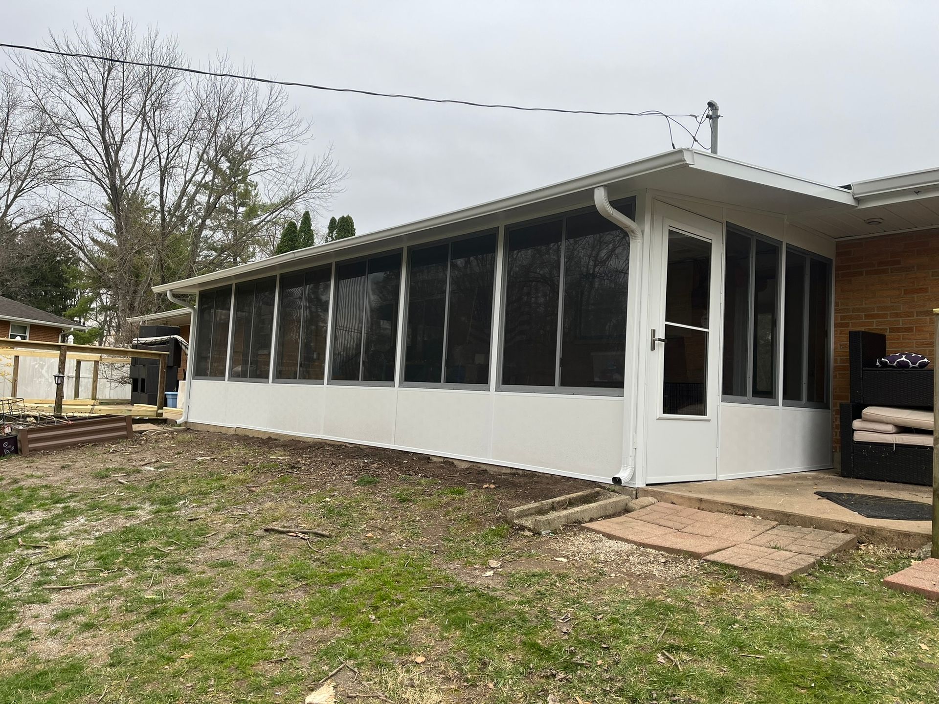 White screened-in porch attached to a brick house, overlooking a yard with some grass and brick pavers.
