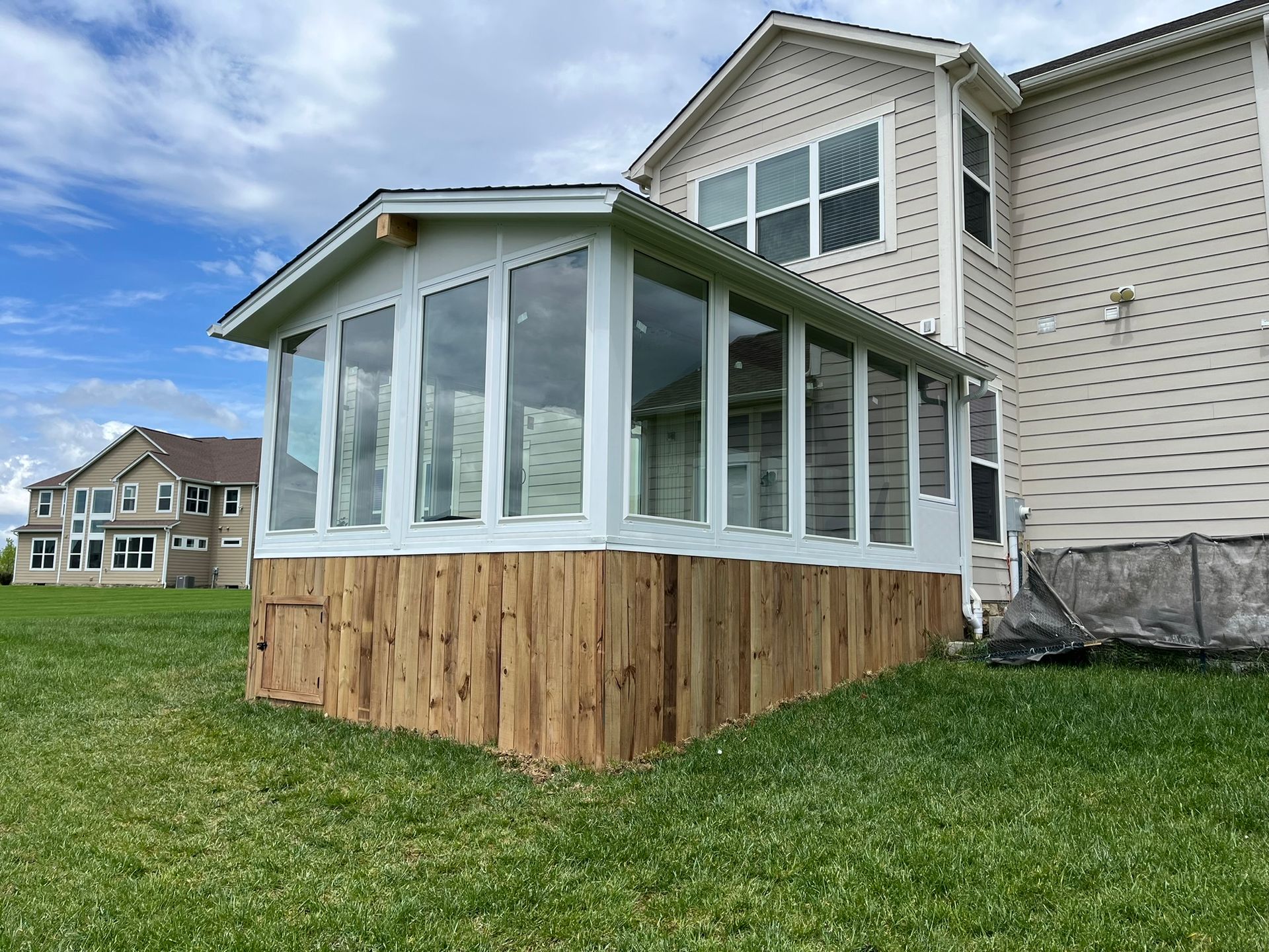 Sunroom addition to a house with white windows and siding, built on a wooden base, set on green grass.