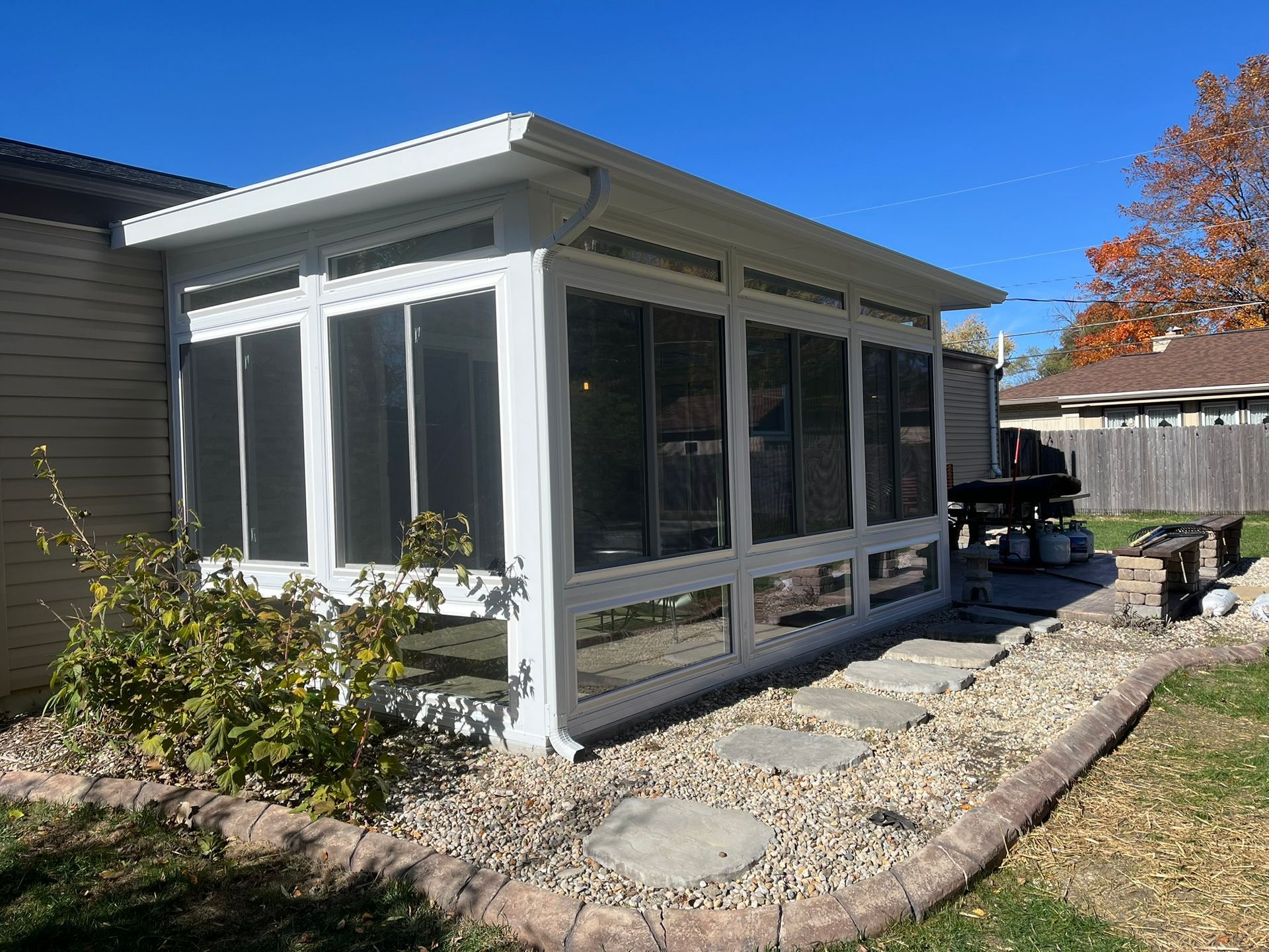 White screened-in porch attached to a beige house, stone walkway, rock garden, and wooden bench.