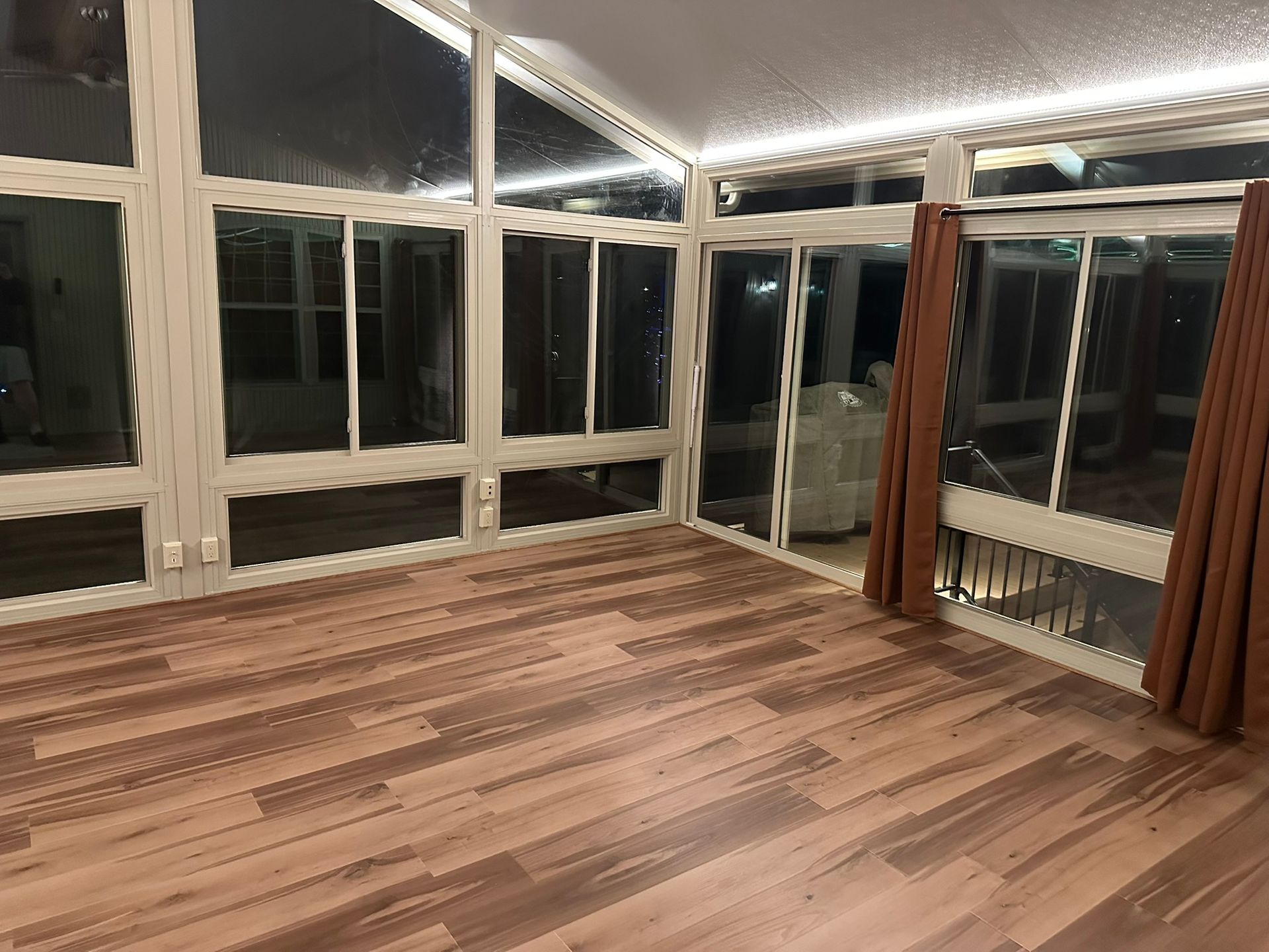 Empty sunroom with glass walls, brown curtains, and wood-look flooring.
