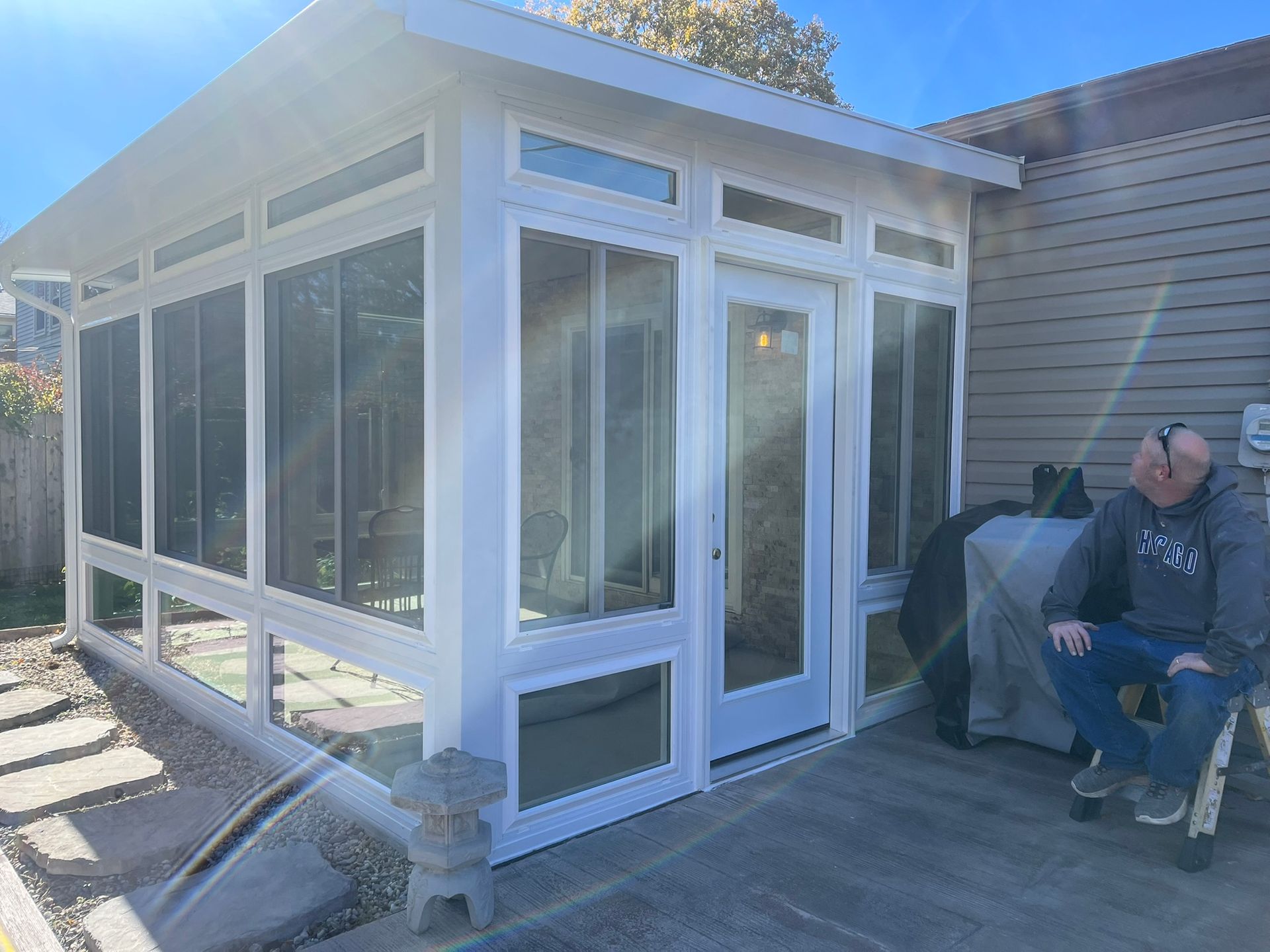 White sunroom with glass windows and a door, attached to a house. Man seated nearby, looking at the room.