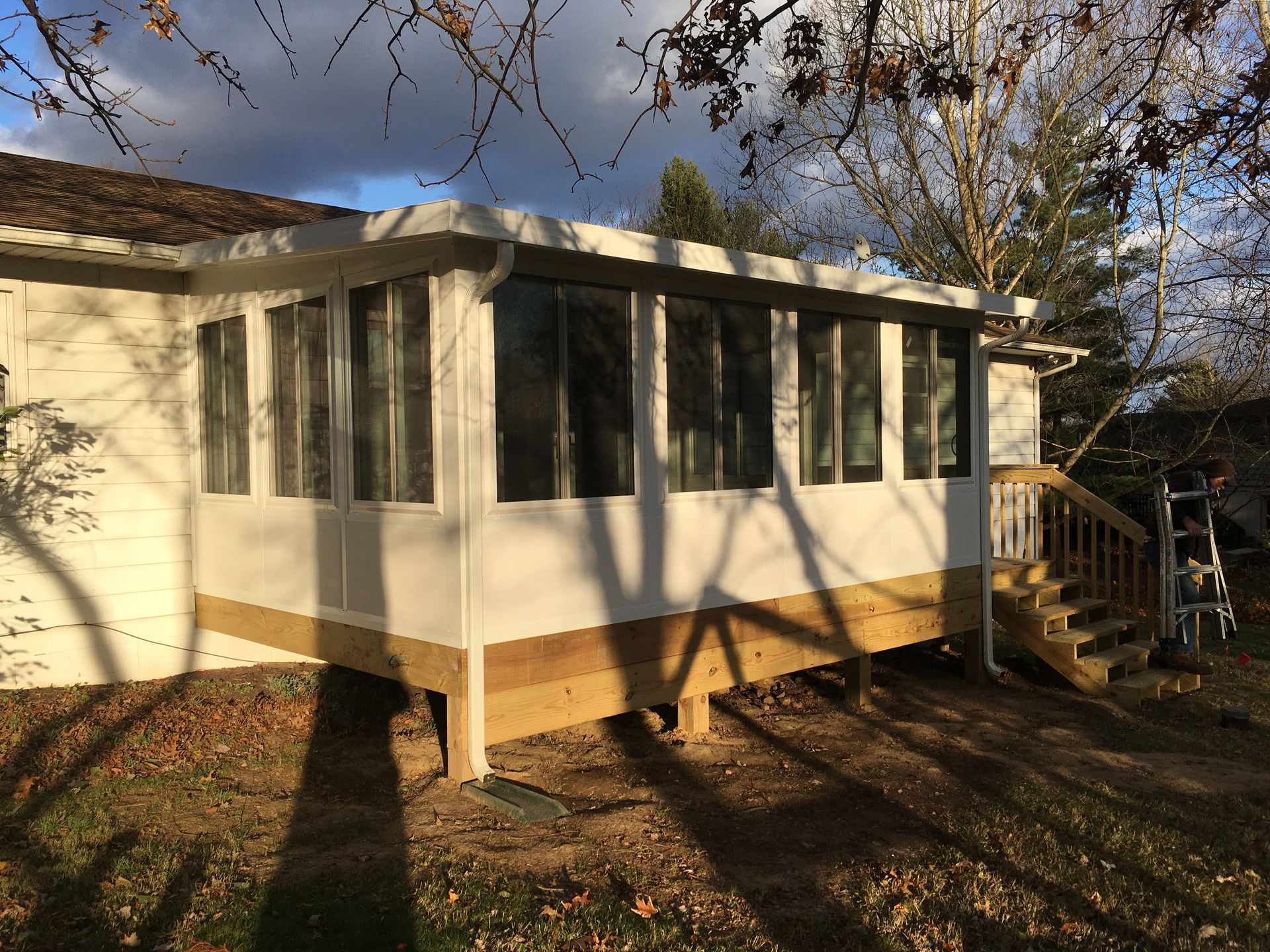 Sunroom addition on a house with a wooden deck, windows, and a dark brown roof. Autumn leaves on the ground.