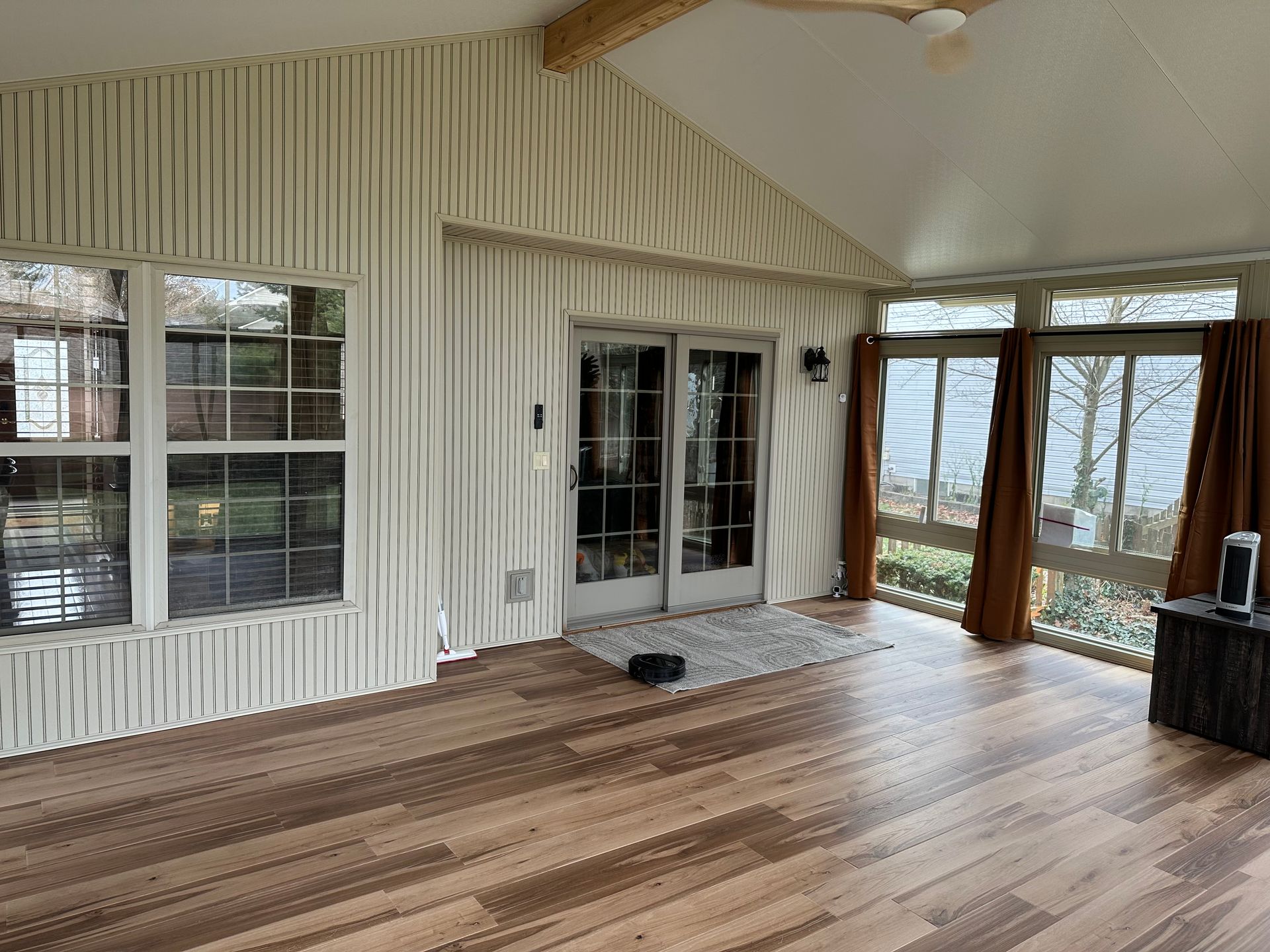 Sunroom with wood floors, windows, and glass doors, with textured vertical wall panels and brown curtains.