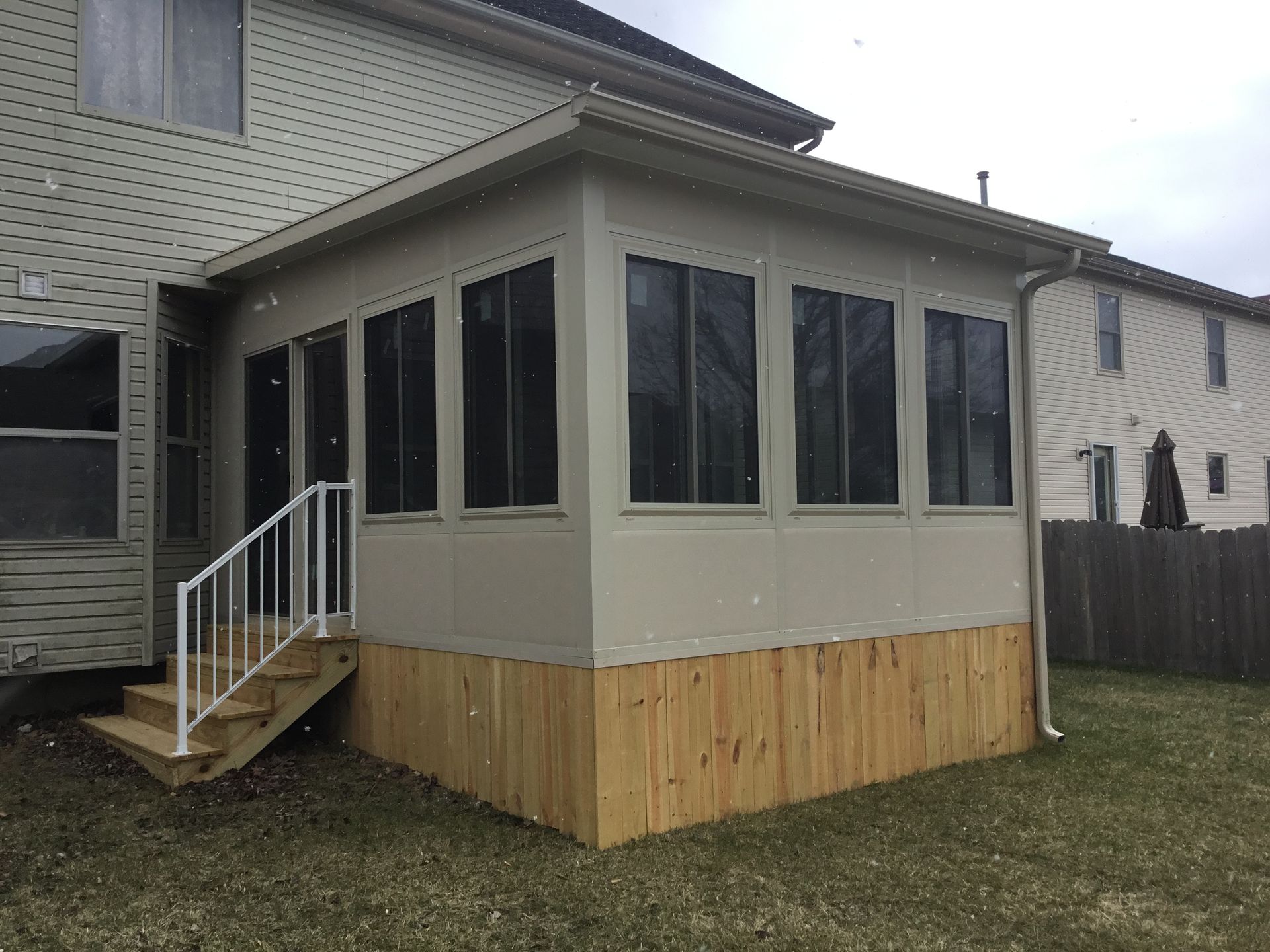 Beige sunroom addition with wooden base, attached to a light-colored house. Stairs with white railing.