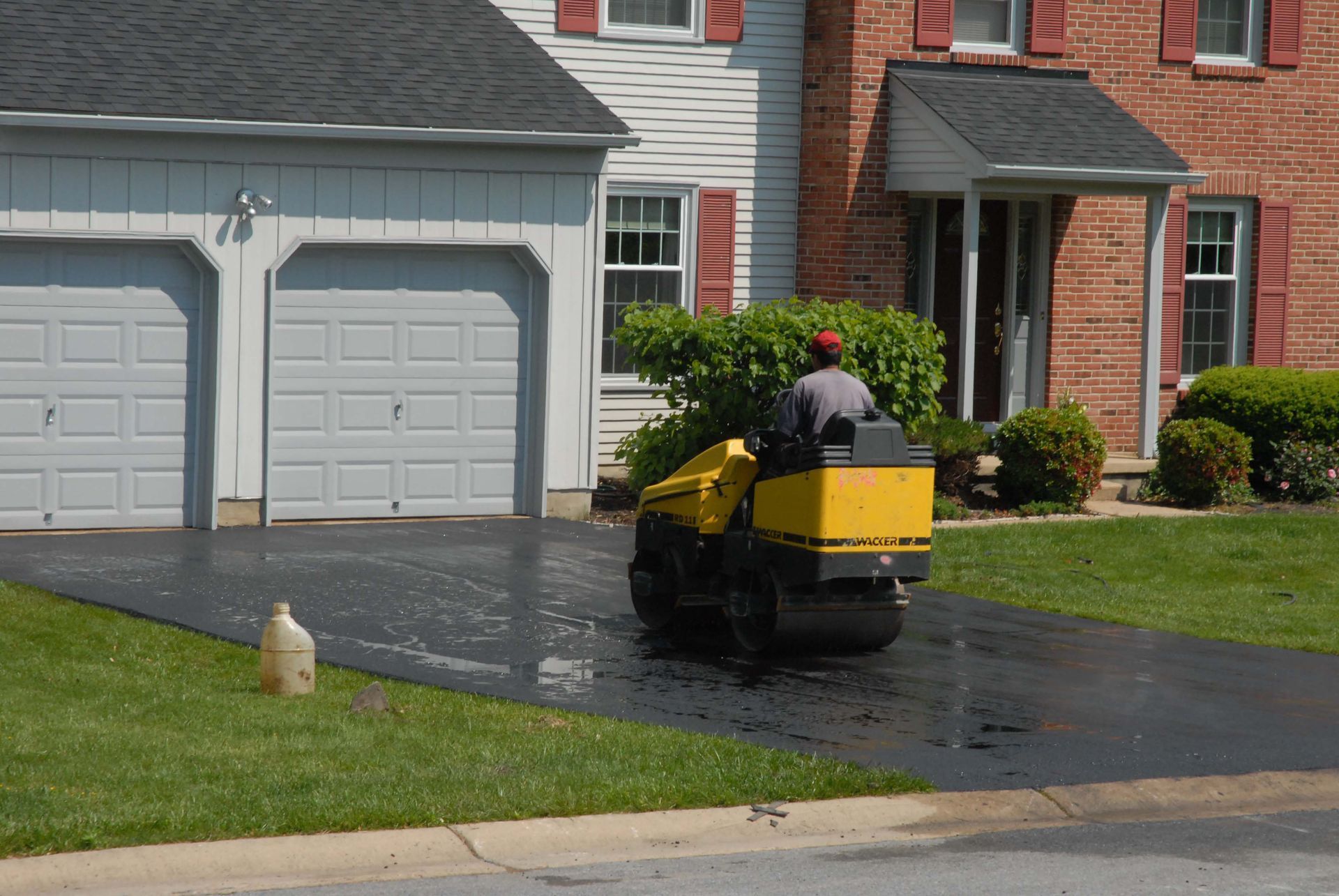 A person operating a yellow asphalt roller compacting a newly paved driveway in front of a house.