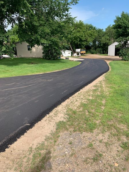 New asphalt driveway curves through a grassy yard, leading to houses and trees under a sunny sky.