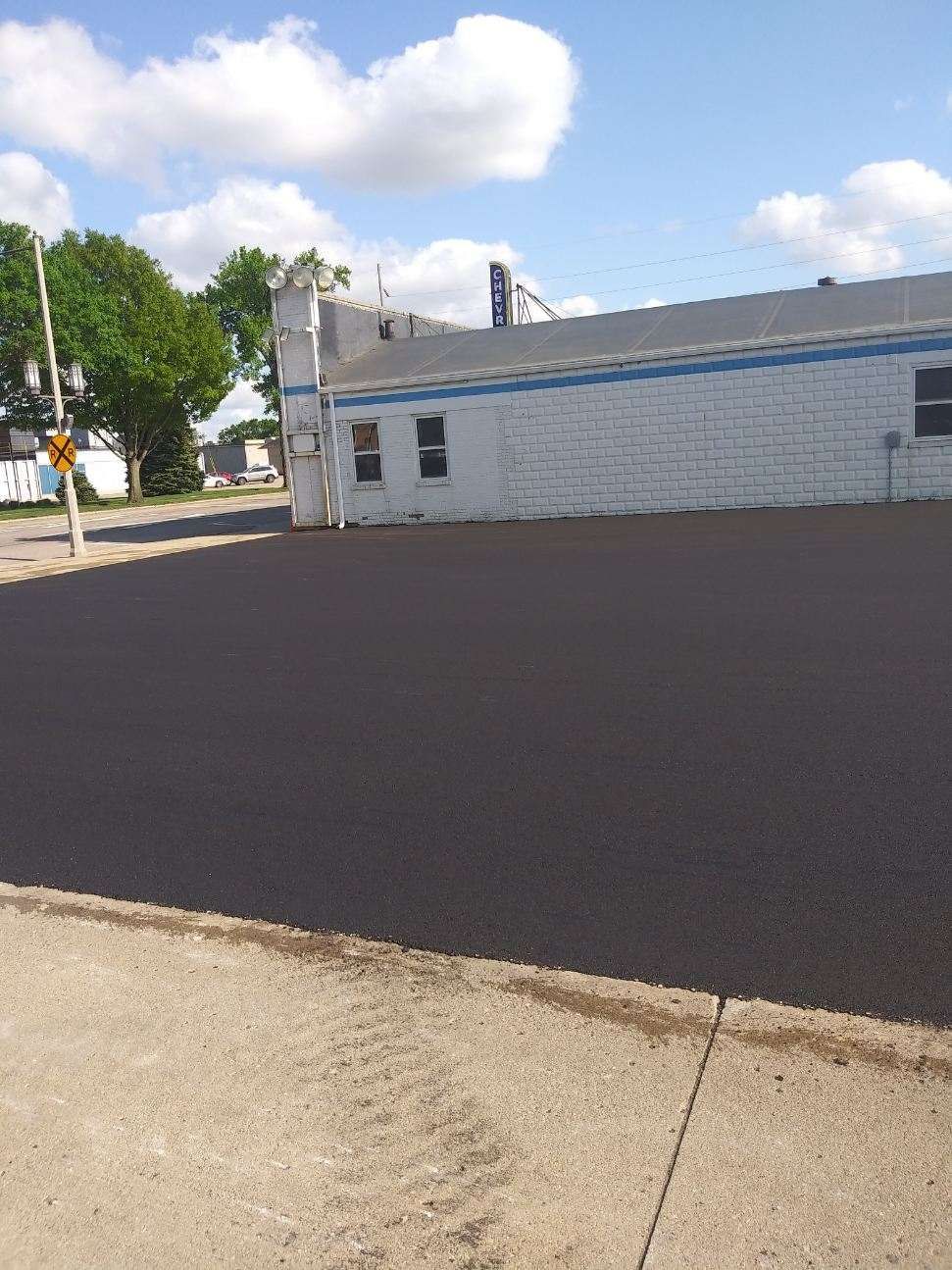 New asphalt parking lot in front of a white building with blue trim. Blue sky and some trees.