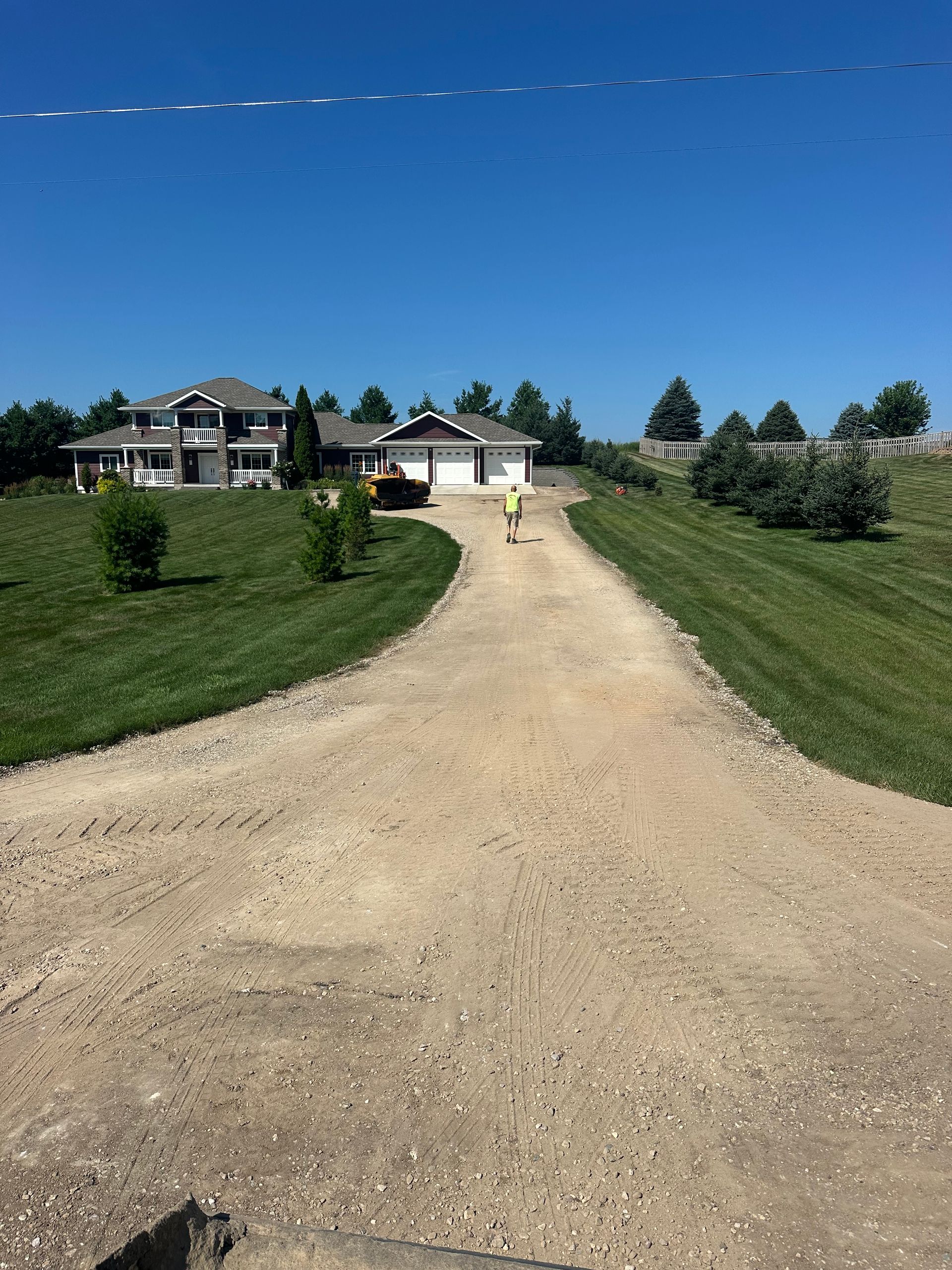 Gravel driveway leading to a house with a person walking towards the garage under a clear, blue sky.