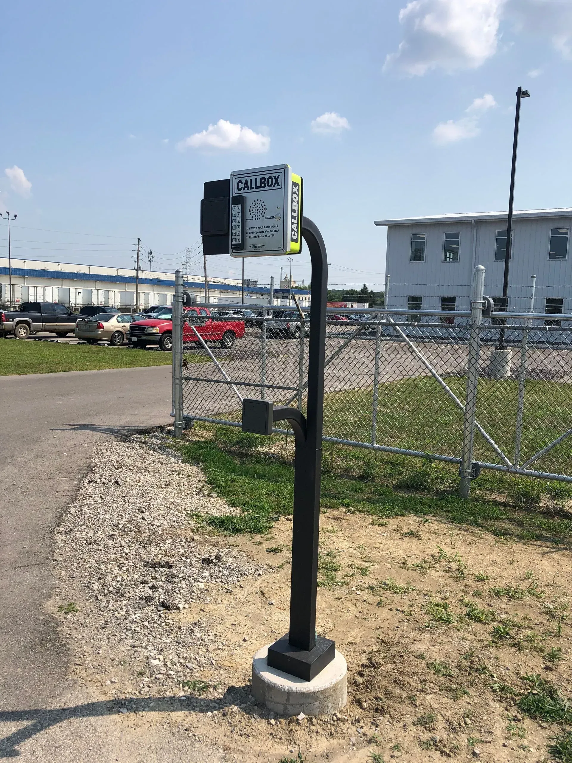 A parking meter is sitting next to a fence in a parking lot.